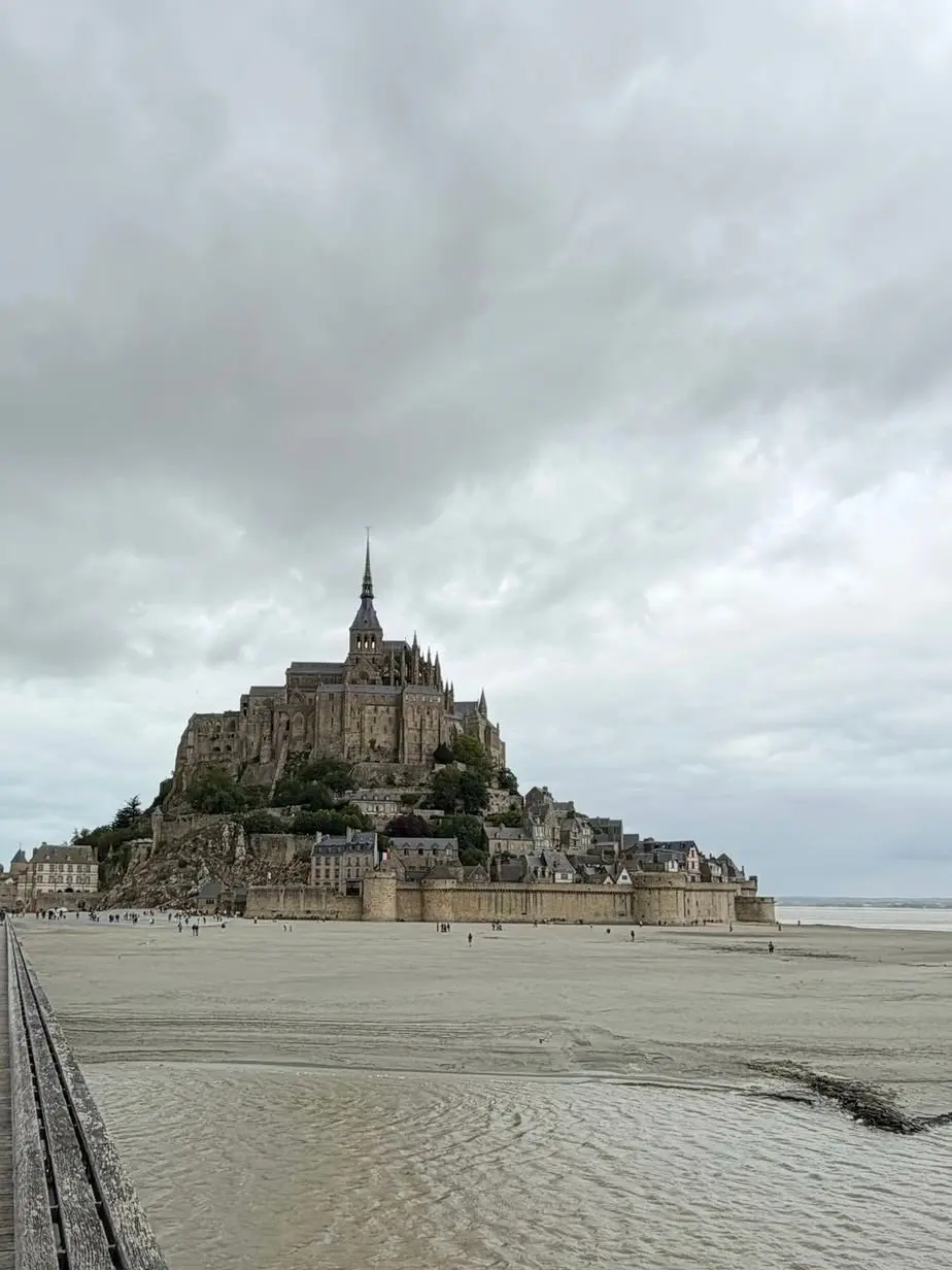 フランス、モン・サン＝ミシェル。岩場からそびえ立つ中世の修道院と集落。砂浜が広がり、曇り空が背景に広がる。