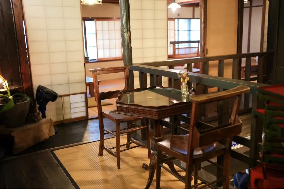 木製の椅子とテーブルが置かれた、日本の伝統的な部屋の様子。畳敷きの床に、アンティーク調の家具が配置されている。背景には障子や窓が見える。
