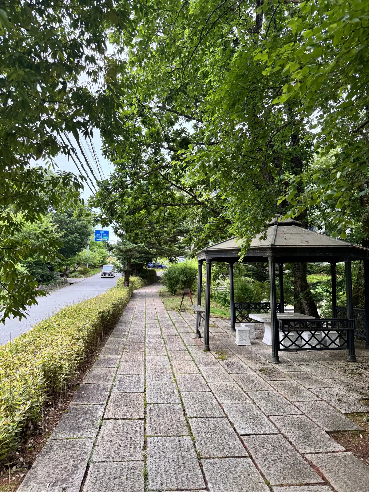 石畳の道が、緑豊かな木々の下を通って遠くの休憩所に続いています。道の両脇には低木が植えられ、休憩所は黒い柱と屋根で構成されています。背景には、道路と車が見えます。
