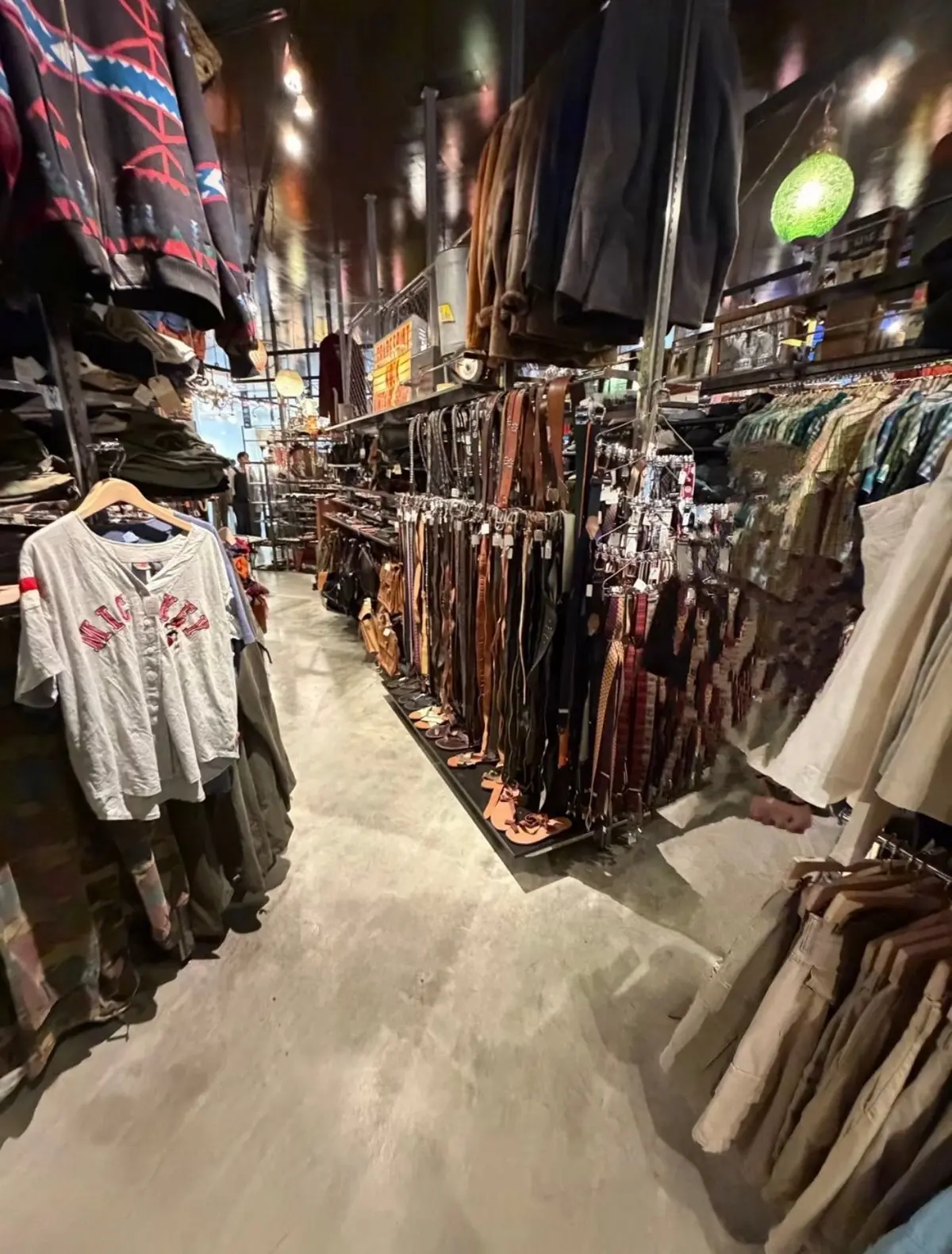 古着屋の店内。中央にはベルトが並び、左右に服が掛けられている。左にはTシャツ、右にはジャケットやワンピースなど。天井には照明器具や装飾品が吊り下げられている。