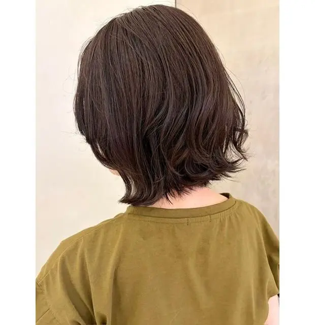 ダークブラウンのショートボブ。肩にかかる長さで、毛先は外側にカールしている。シンプルな緑色のTシャツを着用。