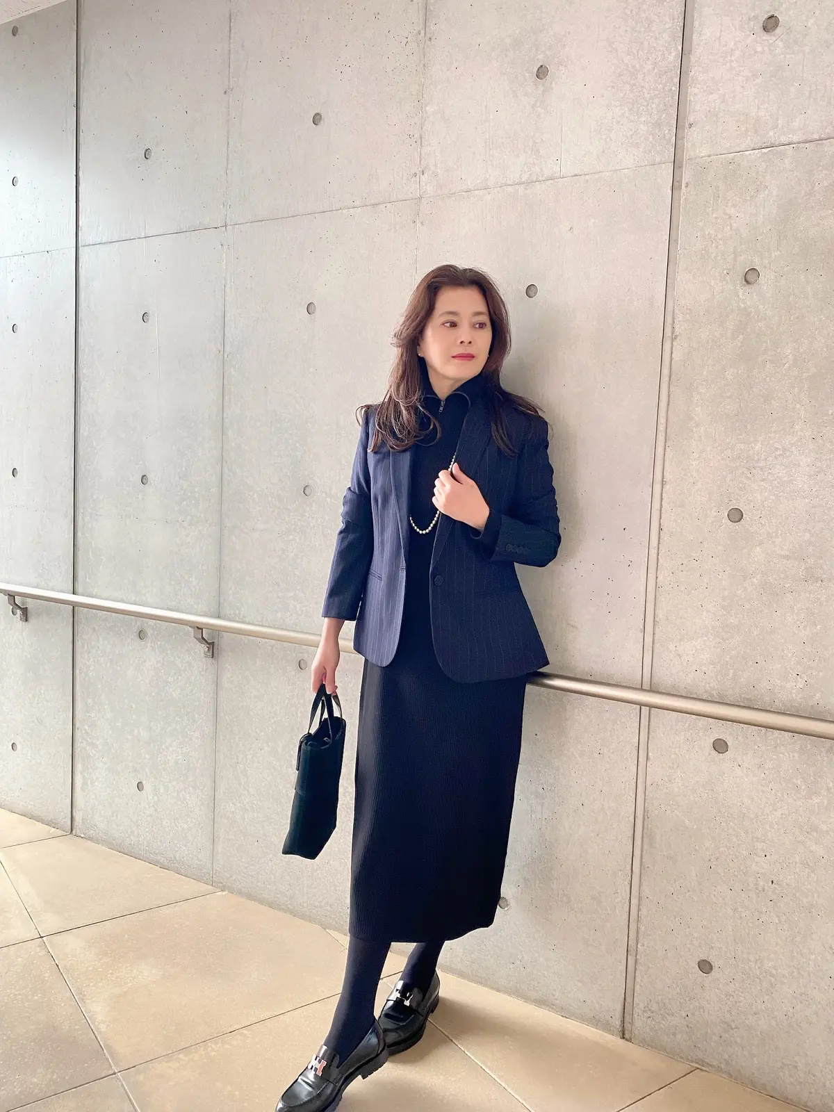 コンクリートの壁にもたれ黒のユニクロハーフジップニットワンピースを着てパールのロングネックレス、ジャケットを羽織り横を向いた女性の写真