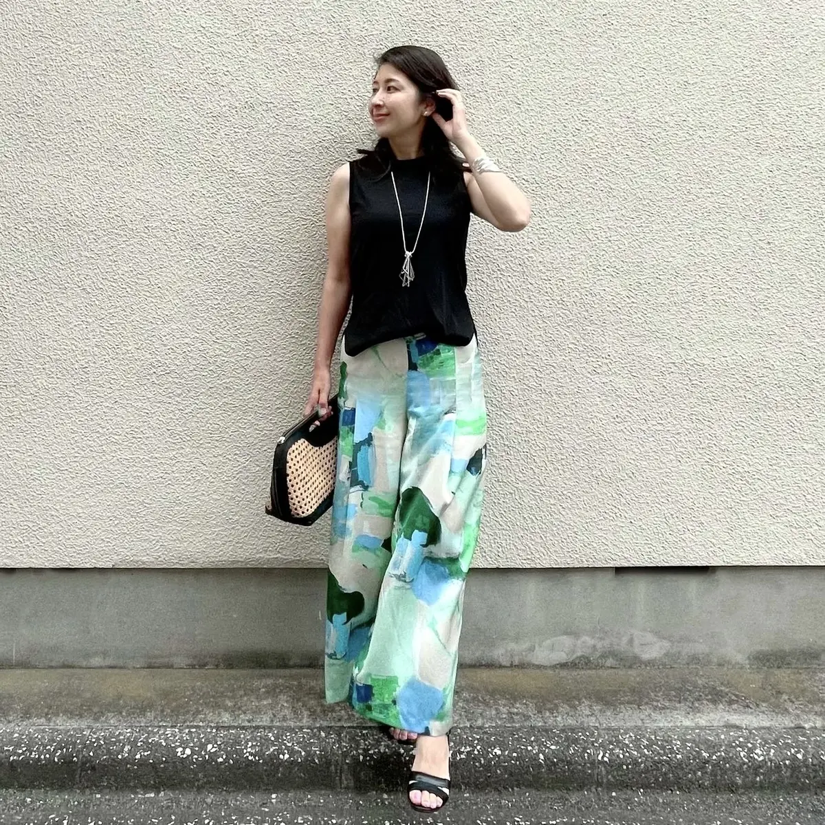黒Tシャツ、ベージュとグリーンのペイント柄パンツ、黒レザーとラフィア素材のハンドバッグ、黒サンダル