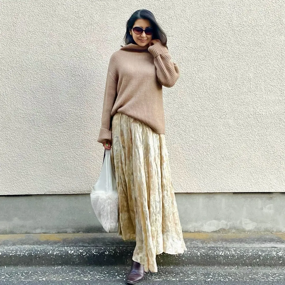 ベージュのローゲージニット、ゴールドの花柄スカート、白のファーバッグ、茶のロングブーツ