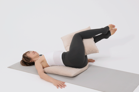 OurAge×Webエクラ　クッションのひとつはお尻の下に、ひとつは膝を曲げて、内ももで挟みます。腰を床に押しつけるようにして足を膝の高さに上げて。息を吐きながらゆっくり両脚を倒します。両肩が床から離れる直前で止め、息を吸いながらゆっくり元へ戻りましょう。左右交互に10回ずつ。