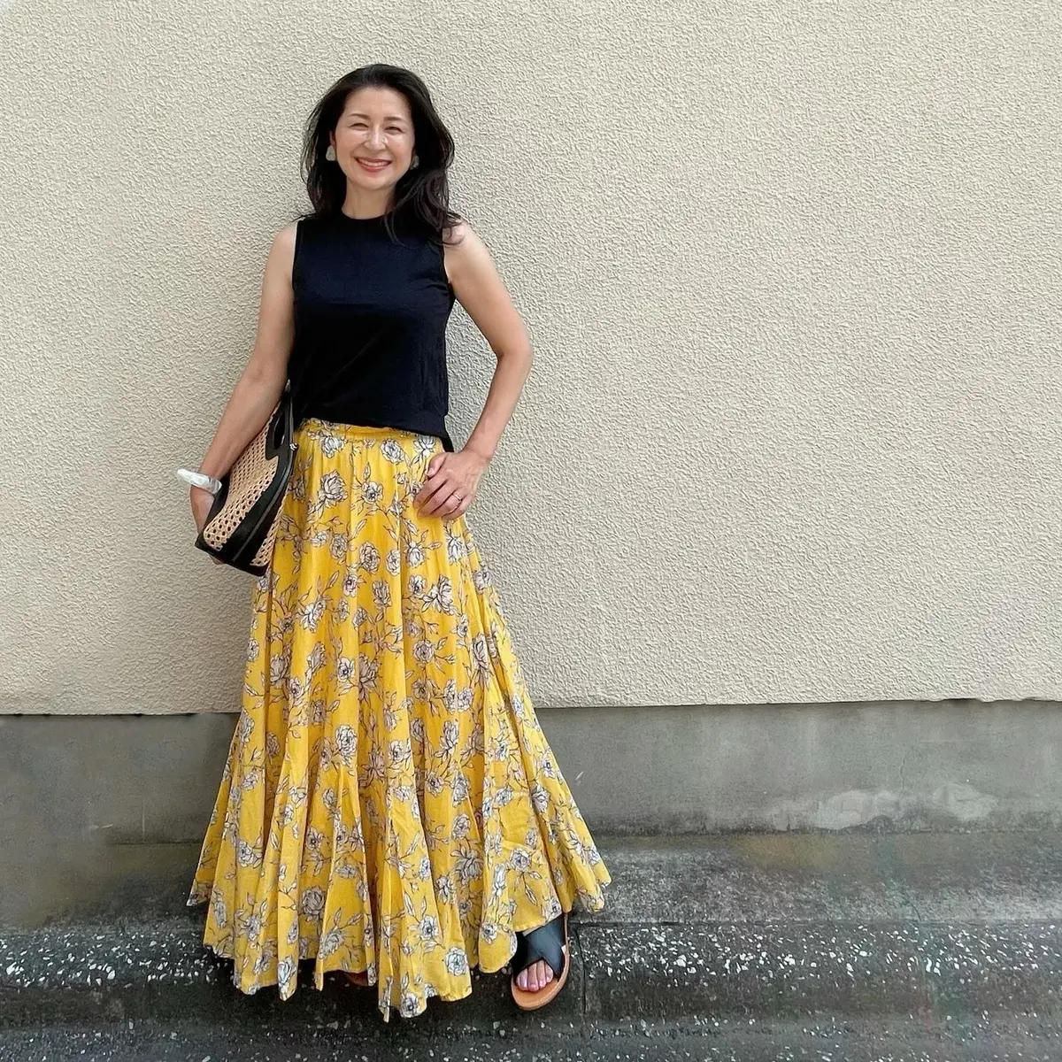 黒ノースリーブTシャツ、イエローの花柄スカート、黒フラットサンダル、黒のレザーとラフィアのバッグ