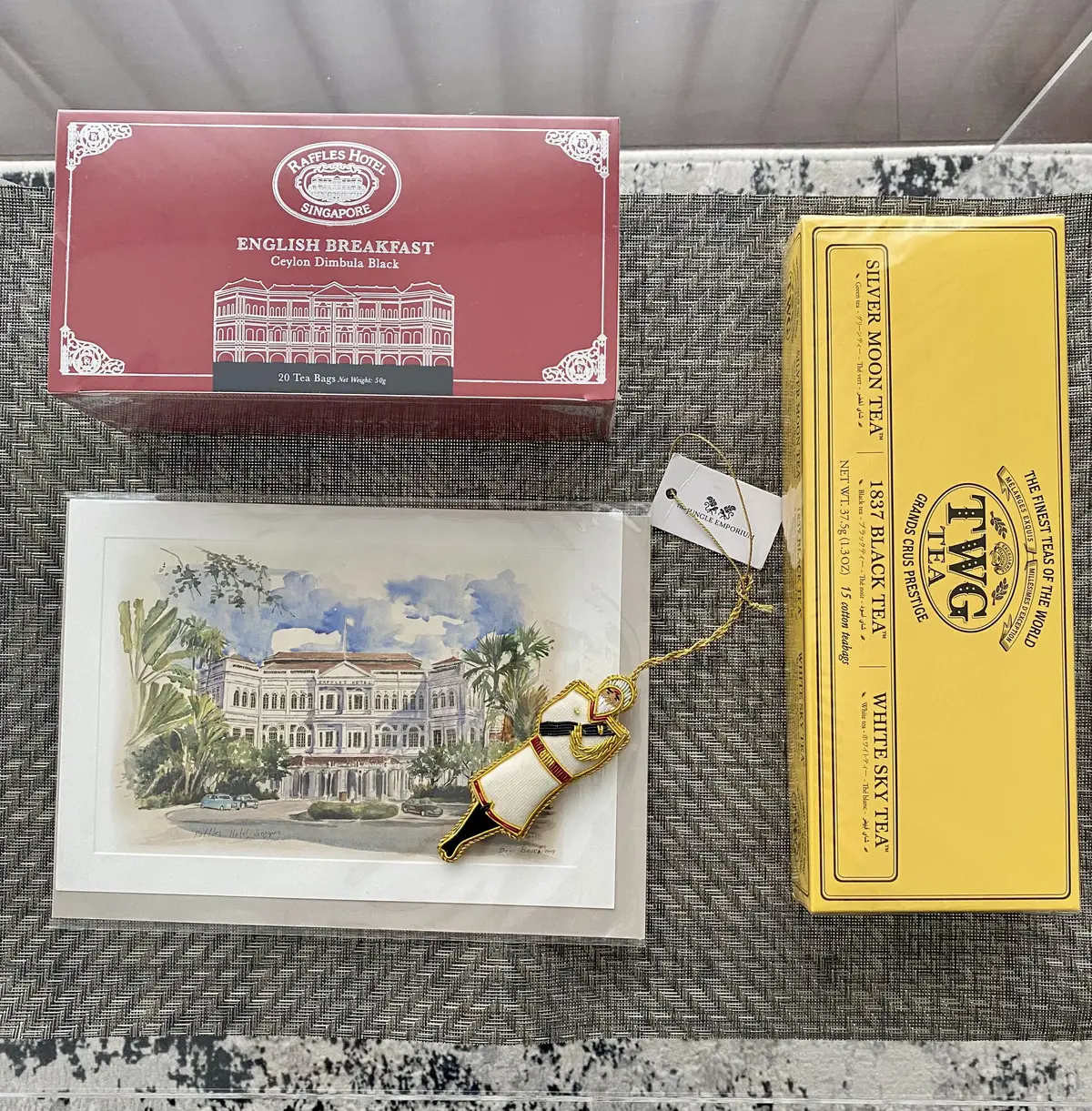 ラッフルズホテルの紅茶とTWGのティーバッグの詰め合わせ、水彩のポストカード、ラッフルズの制服を模したオーナメントをテーブルに並べたお土産写真。