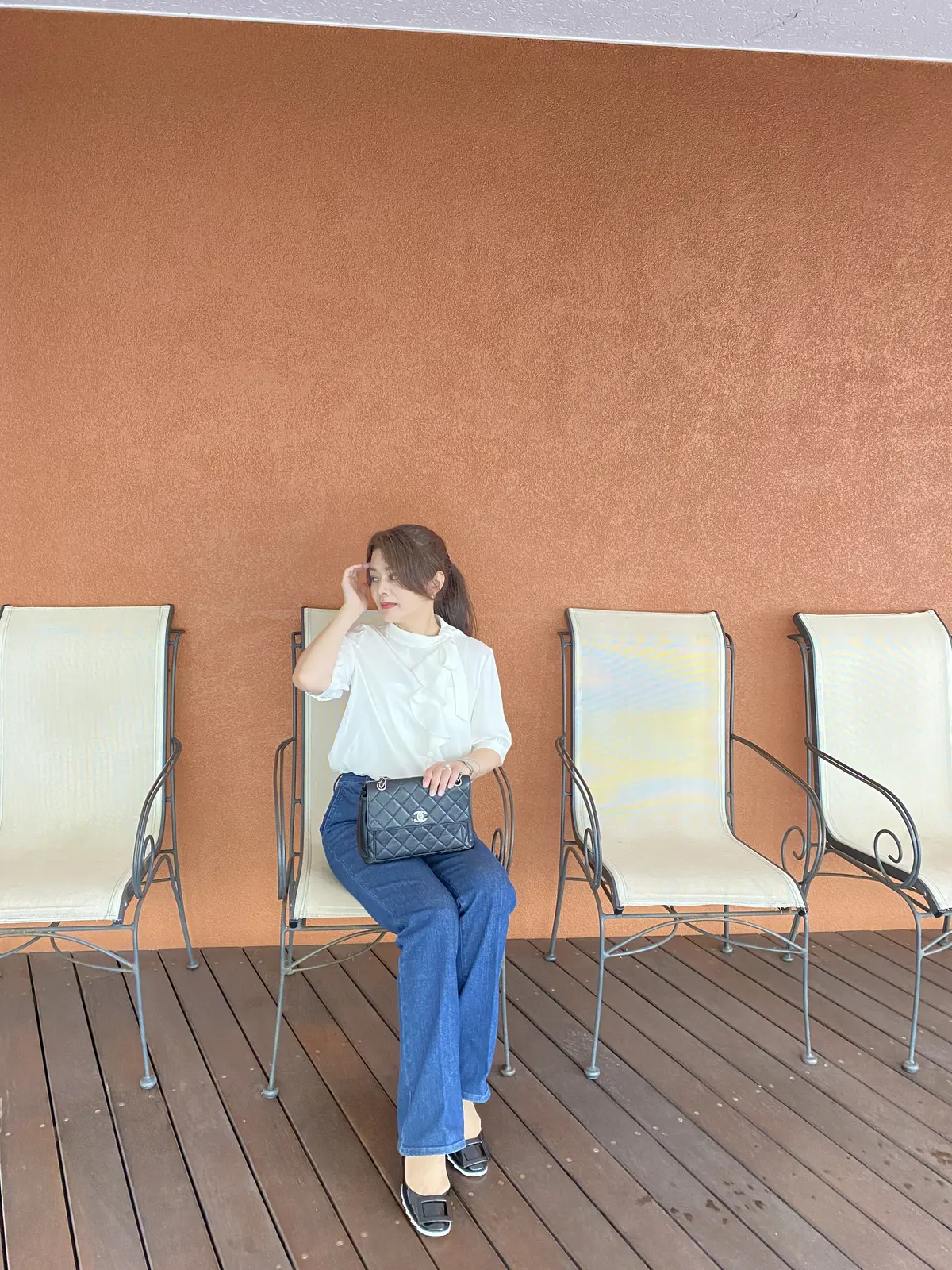 五分袖巻かないボウタイブラウス〈オフホワイト〉と、スリムフレアトラウザージーンズ〈ネイビー〉を着た女性が、テラスのアイアンチェアに座り、シャネルのチェーンバッグを膝に置いて微笑む写真。足元は黒のフラットシューズでまとめ、背景のテラコッタカラーの壁とウッドデッキが、コーディネートを引き立てている。
