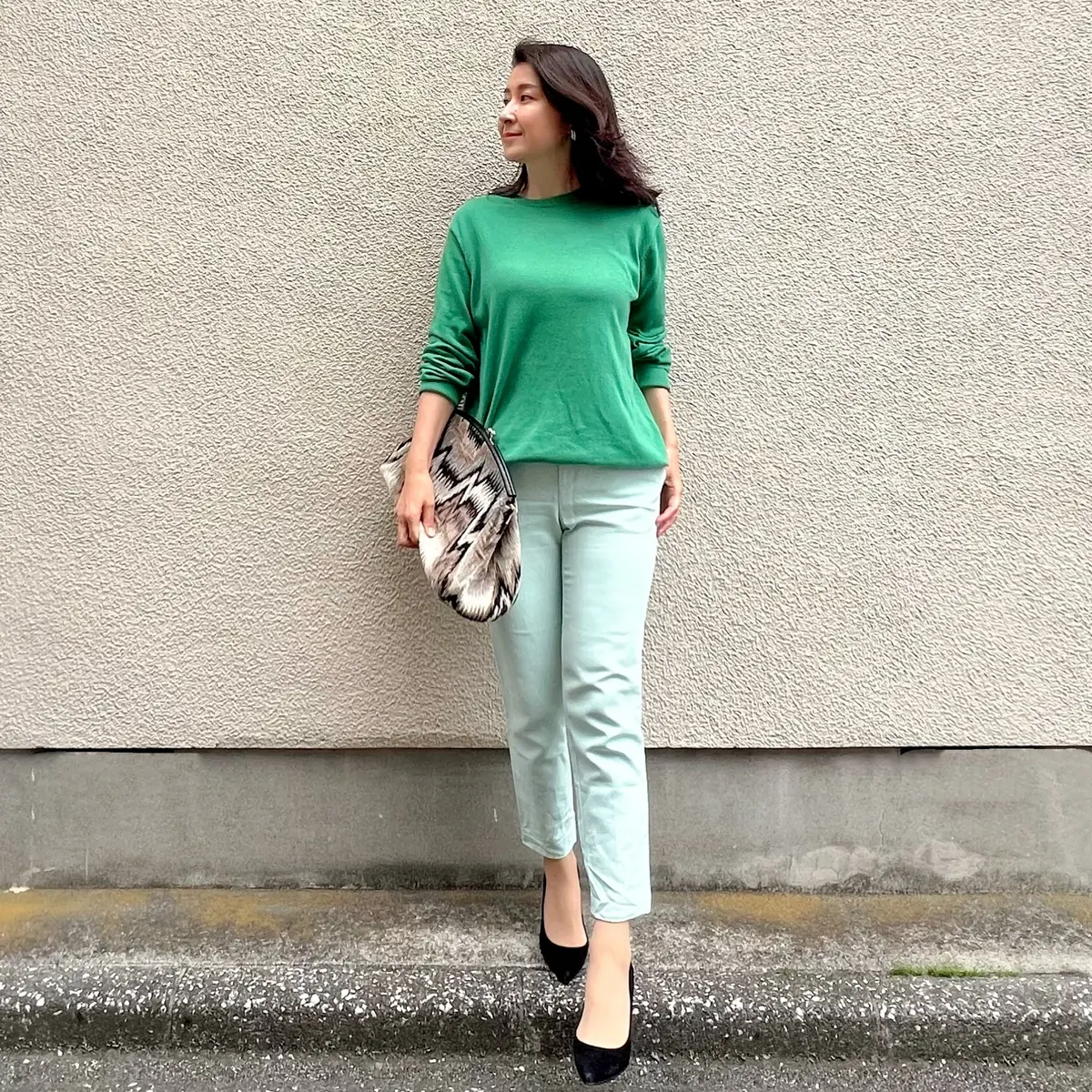 グリーンのニット、薄めグリーン色のパンツ、黒系の柄物のクラッチバッグ、黒のパンプス