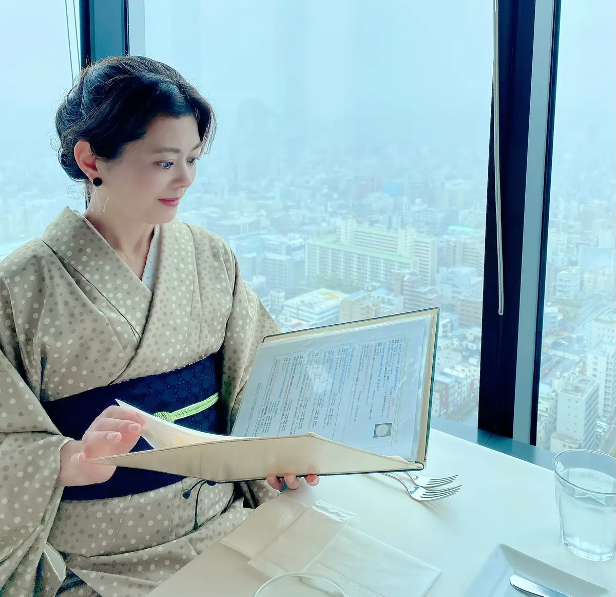 ブラッスリーオザミ東京スカイツリーソラマチ店のスカイツリーの見える窓際席に座りメニューを眺める和服姿の女性の画像