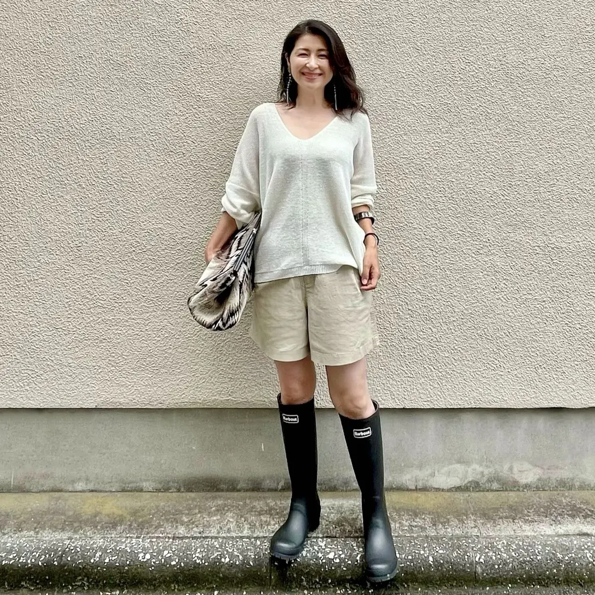 白のサマーニット、ベージュのショートパンツ、黒のロングレインブーツ、大きめクラッチバッグ