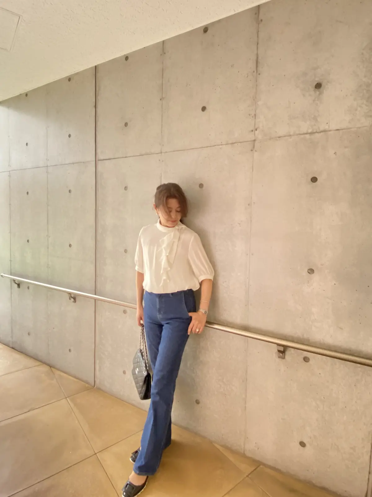 五分袖ボウタイブラウス〈オフホワイト〉とスリムフレアトラウザージーンズ〈ネイビー〉を着た女性が、コンクリート壁の前に立ち、足元にはロジェ ヴィヴィエの黒フラットシューズとシャネルのチェーンバッグを合わせた全身コーディネート写真。