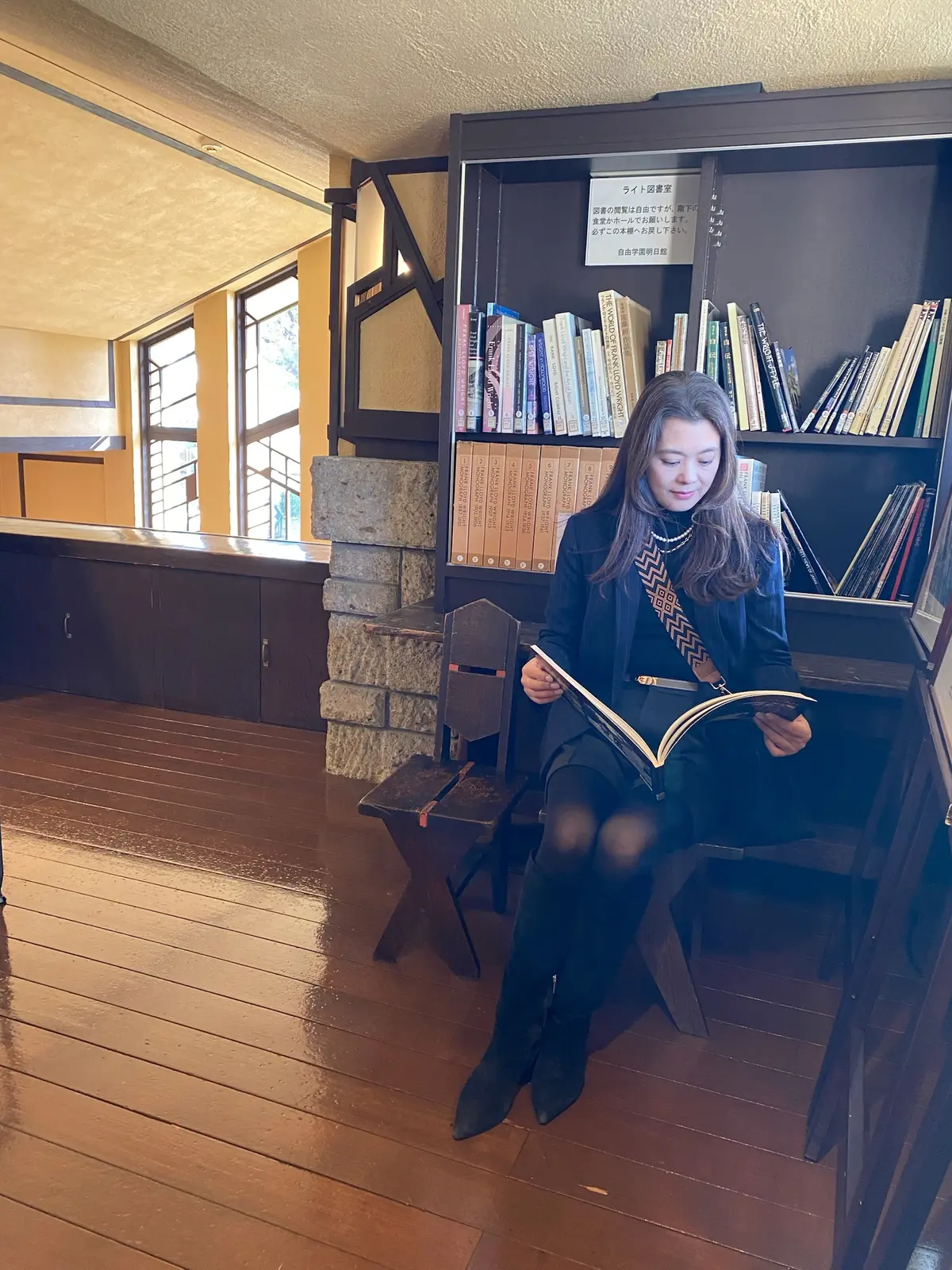 フランク・ロイド・ライト設計の自由学園明日館の図書室で、本を読む女性。木の床と書架、自然光が印象的な落ち着いた空間。