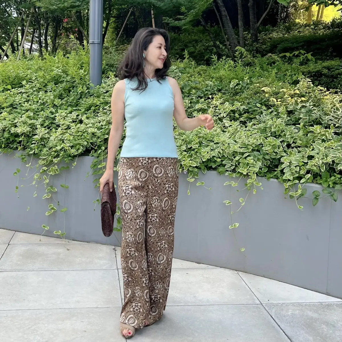 柄物ワイドストレートパンツ、ライトブルーのノースリーブニット、茶のラフィア素材のクラッチバッグ、ベージュのウエッジサンダル
