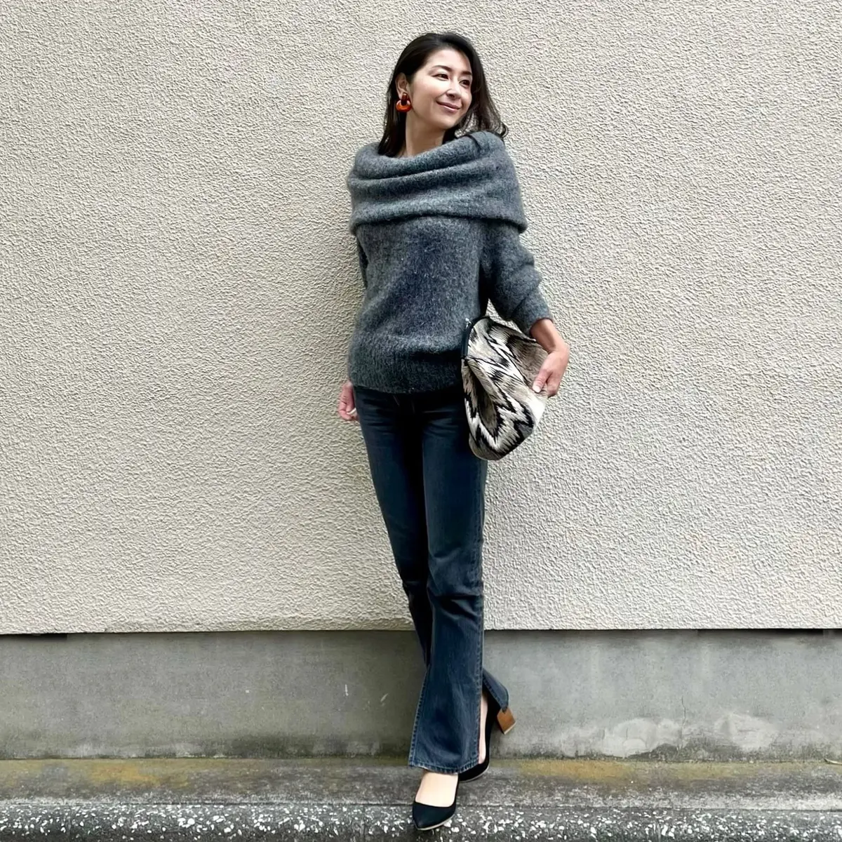 グレーのオフショルダーニット、ブラックのデニム、ブラックのパンプス、大きめのクラッチバッグ、オレンジのイヤリング