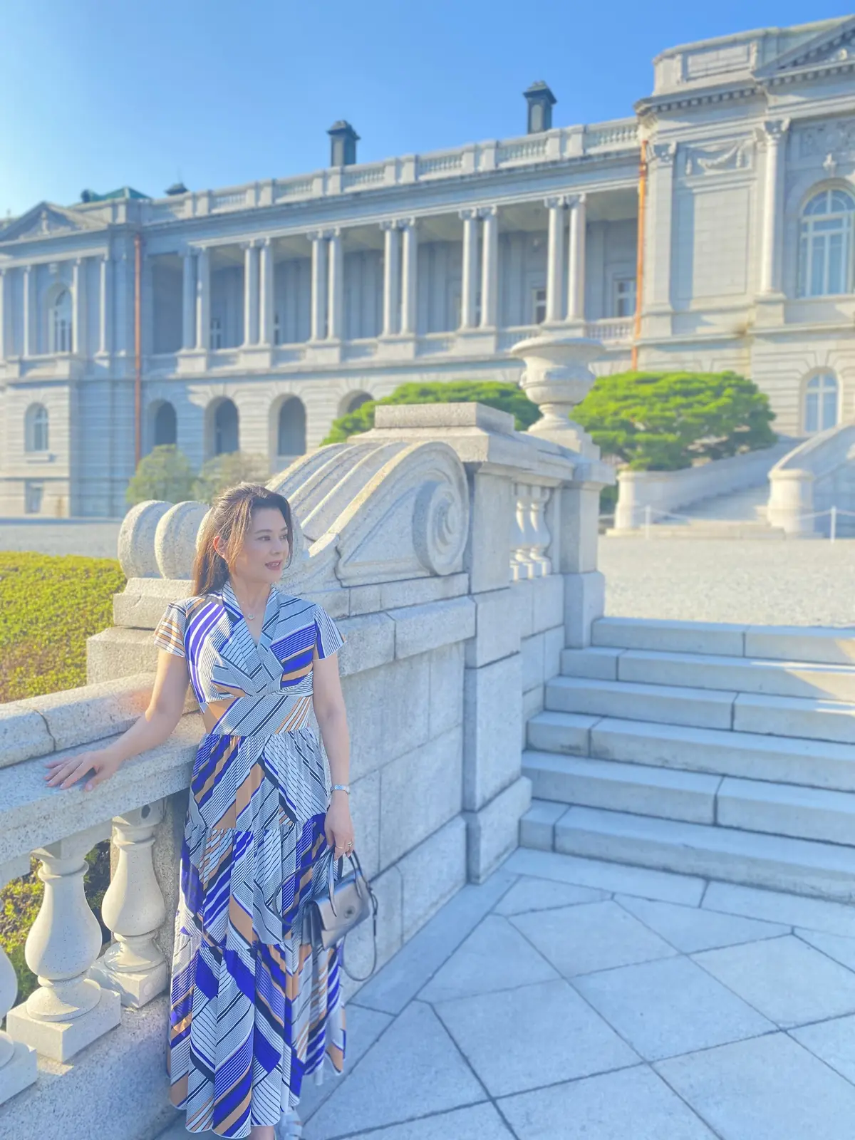 迎賓館赤坂離宮の手すりに寄りかかる女性。ブルーとキャメルの幾何学柄ワンピースが石造りの建物と調和している。