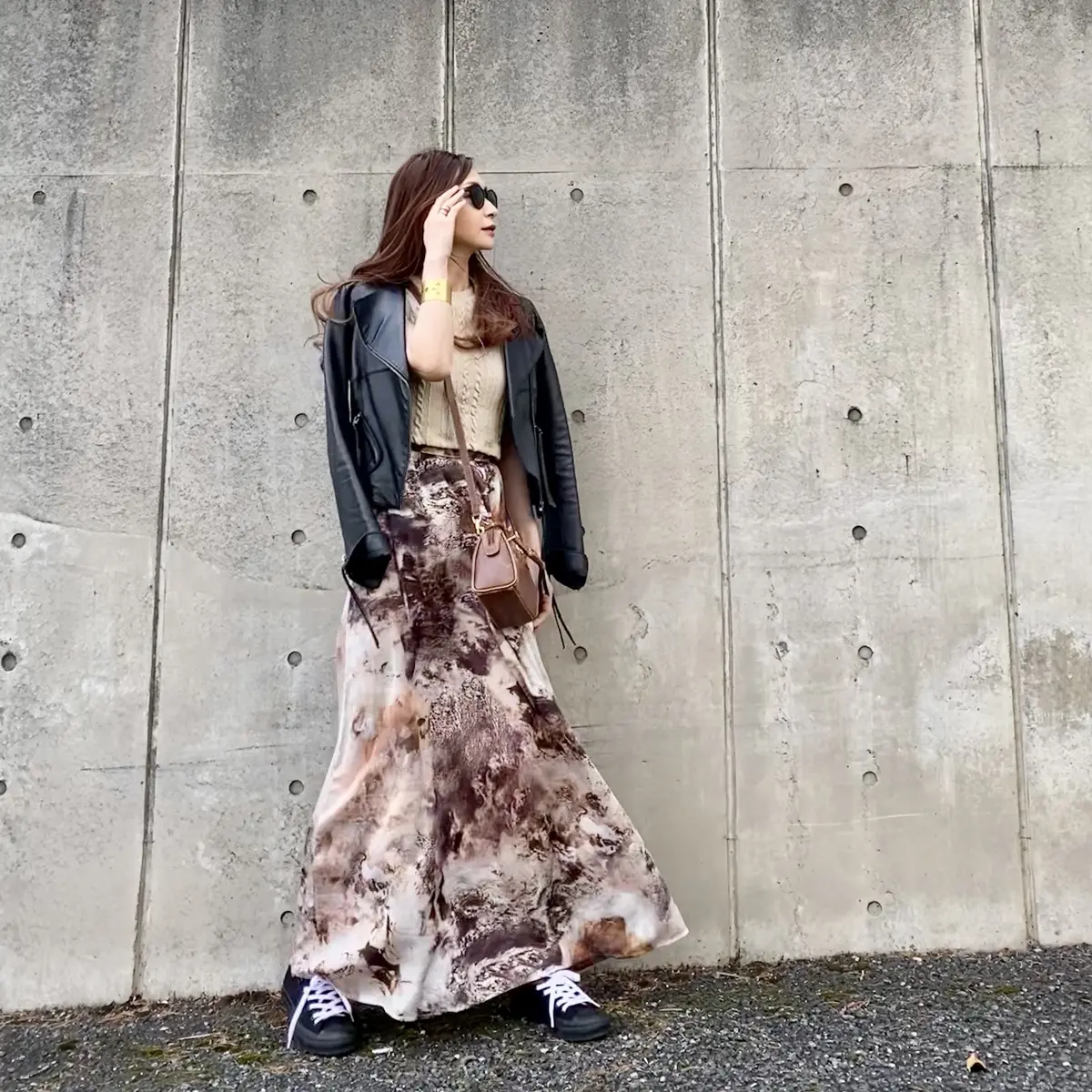 ライダースジャケット、ベージュのショートトップス、ベージュ主体の柄スカートにコンバースのスニーカー