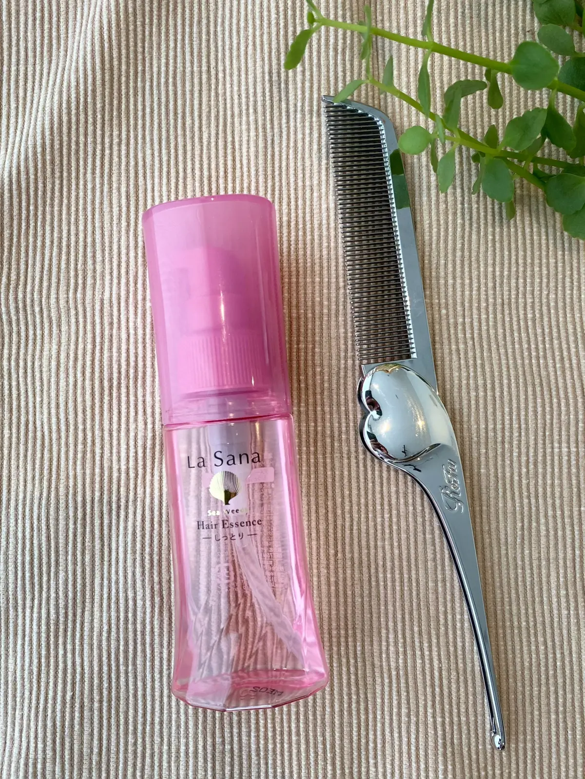 シンプルな布地に置かれたラサーナのピンクのヘアエッセンスボトルと、リファのハートコーム（シルバー）を並べた画像。縮毛矯正の髪にもおすすめのヘアケアアイテム。