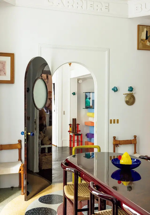 テラゾー床のモチーフと呼応する丸窓のドアが船室を思わせるダイニングルーム。椅子の骨組みと楕円形のテーブルはラムダンがデザインし、フランスきってのラッカー職人に特注した。右手の壁、セラミックのフックにかけた銅羅は日本で購入。食事の用意ができたら、これを叩いて家族に知らせる。テーブルの上のオブジェと、玄関に置いた虹色の棚は、エットーレ・ソットサス