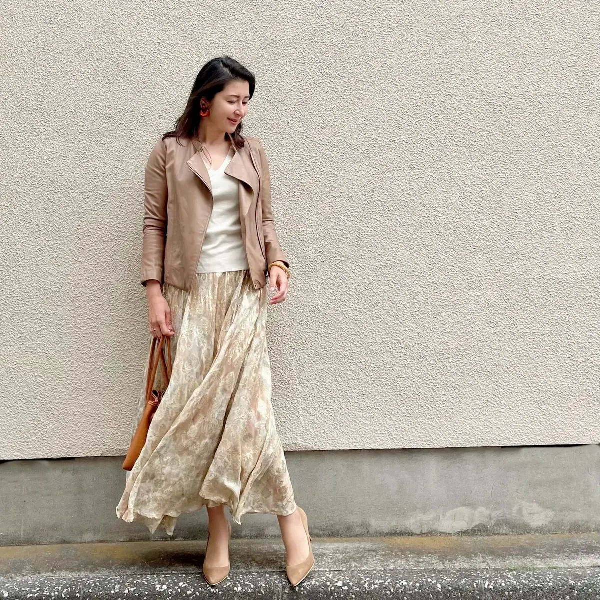 ベージュのライダースジャケット、白のVネックニット、ベージュ系のロングフレアースカート、オレンジ系のバッグ、ベージュのパンプス、オレンジのイヤリング