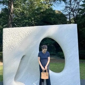 安田侃の大理石彫刻《風》の前でうつむきながら立つ女性。ネイビーの上品な装い。