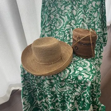 グリーンと白の柄ワンピースを椅子に掛け、ラタン帽子とかごバッグを合わせた南国風コーディネート写真。背景には白いカーテンから柔らかい光が差し込んでいる。