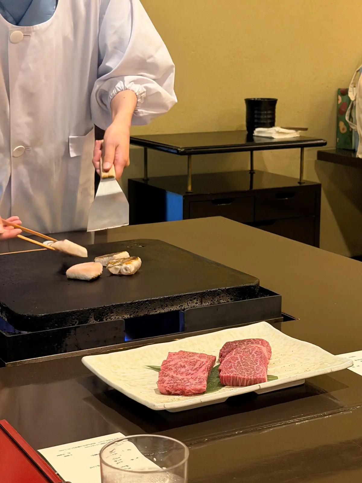 鉄板焼きのテーブルで、白衣を着た調理人が魚を焼いている。手前には和紙の上に並んだ牛肉、奥には黒いキャビネットがある。