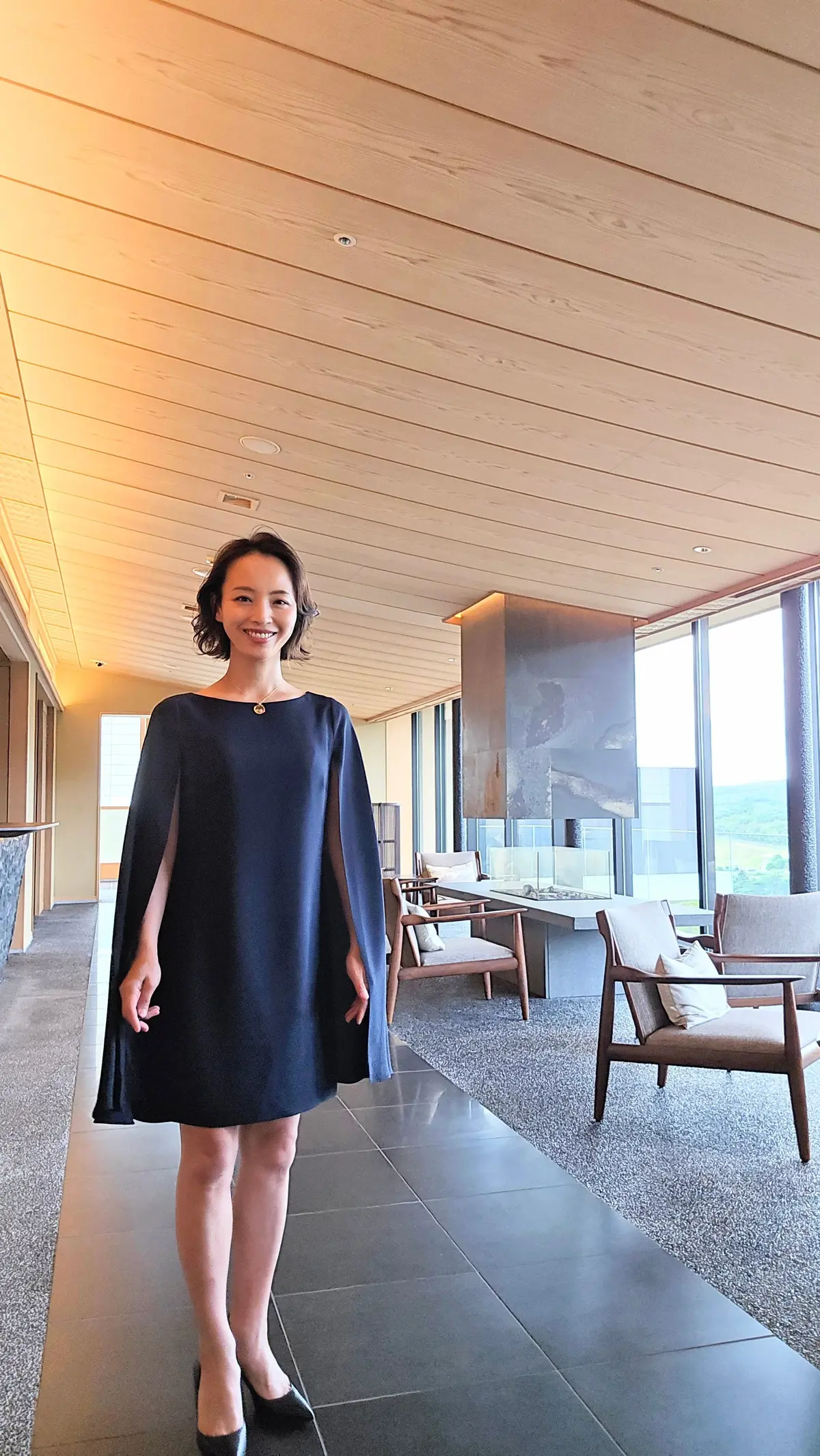 紺色のケープ付きワンピースを着た女性が、明るい空間で微笑んでいる。背景には、木目調の天井、大きな窓、椅子、テーブルが見える。