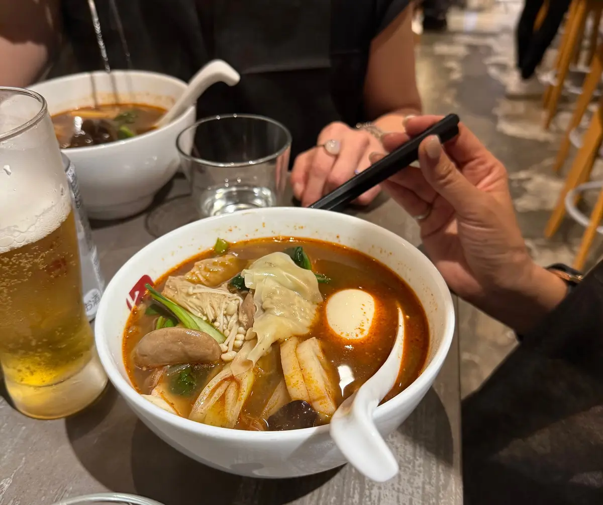 ラーメンの湯気に、アサヒビール、お冷が置かれた食卓。手で箸を持ち、ラーメンを食べる様子。