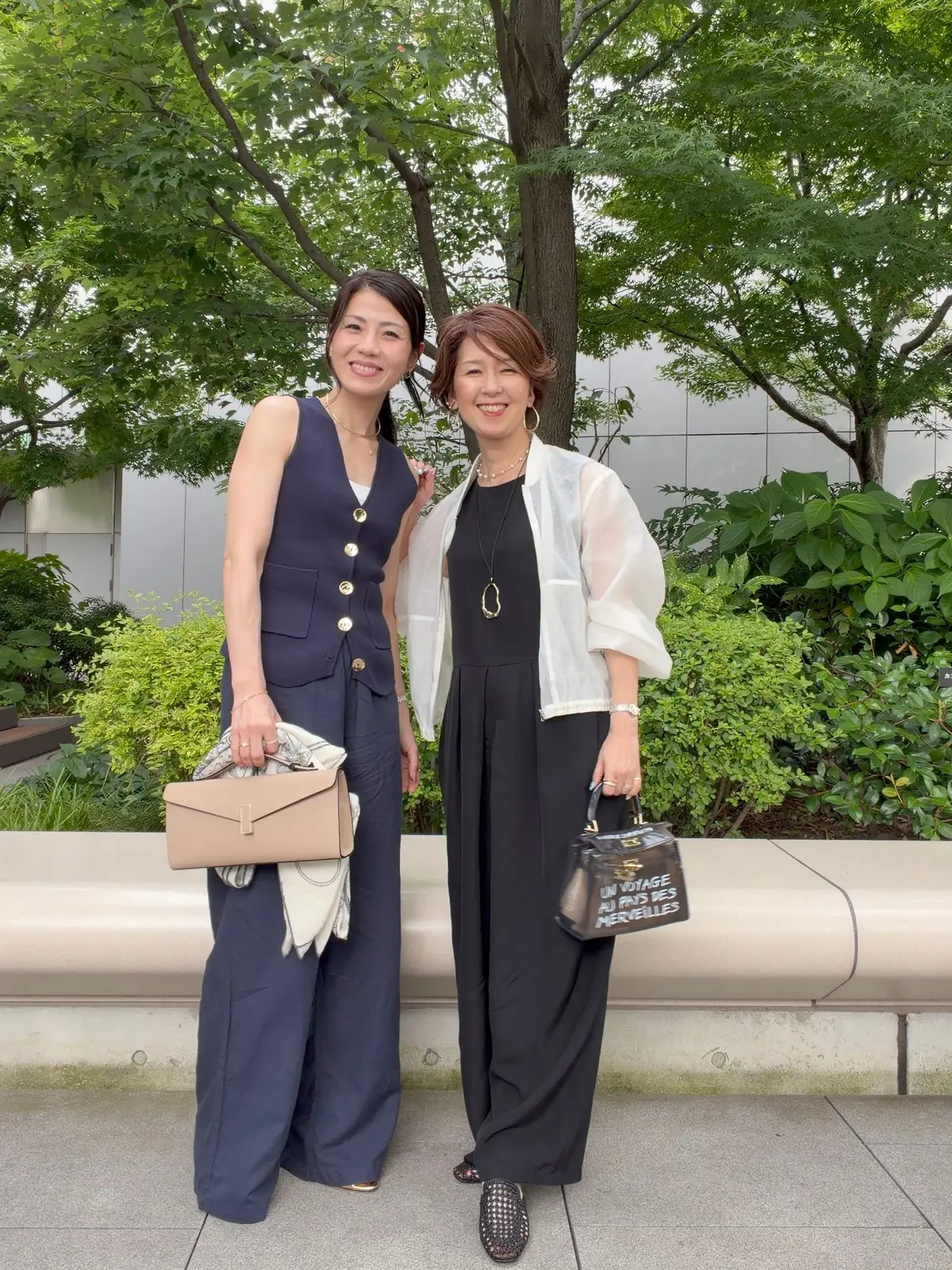二人の女性が屋外で並んで立っている。左の女性は紺色の無地ワンピースにゴールドのボタン、右の女性は黒のワンピースに白のシースルーカーディガン。左の女性はベージュのクラッチバッグ、右の女性は黒のショルダーバッグを所持。背景には緑の木々や低木が広がる。