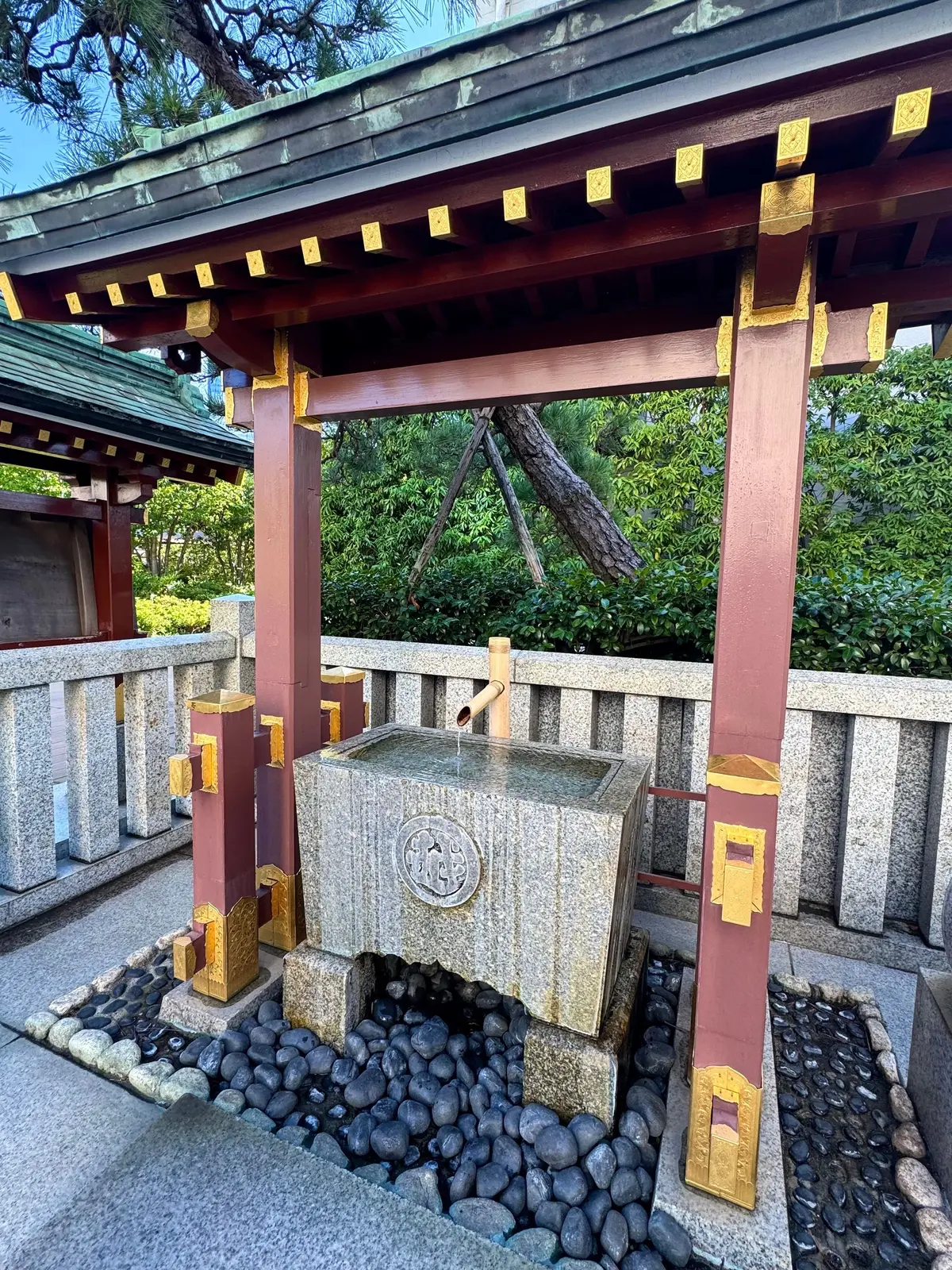 木造の屋根と柱で囲まれた、石造りの手水鉢。黒い小石で敷き詰められた空間に設置され、中央には菊の模様が刻まれている。