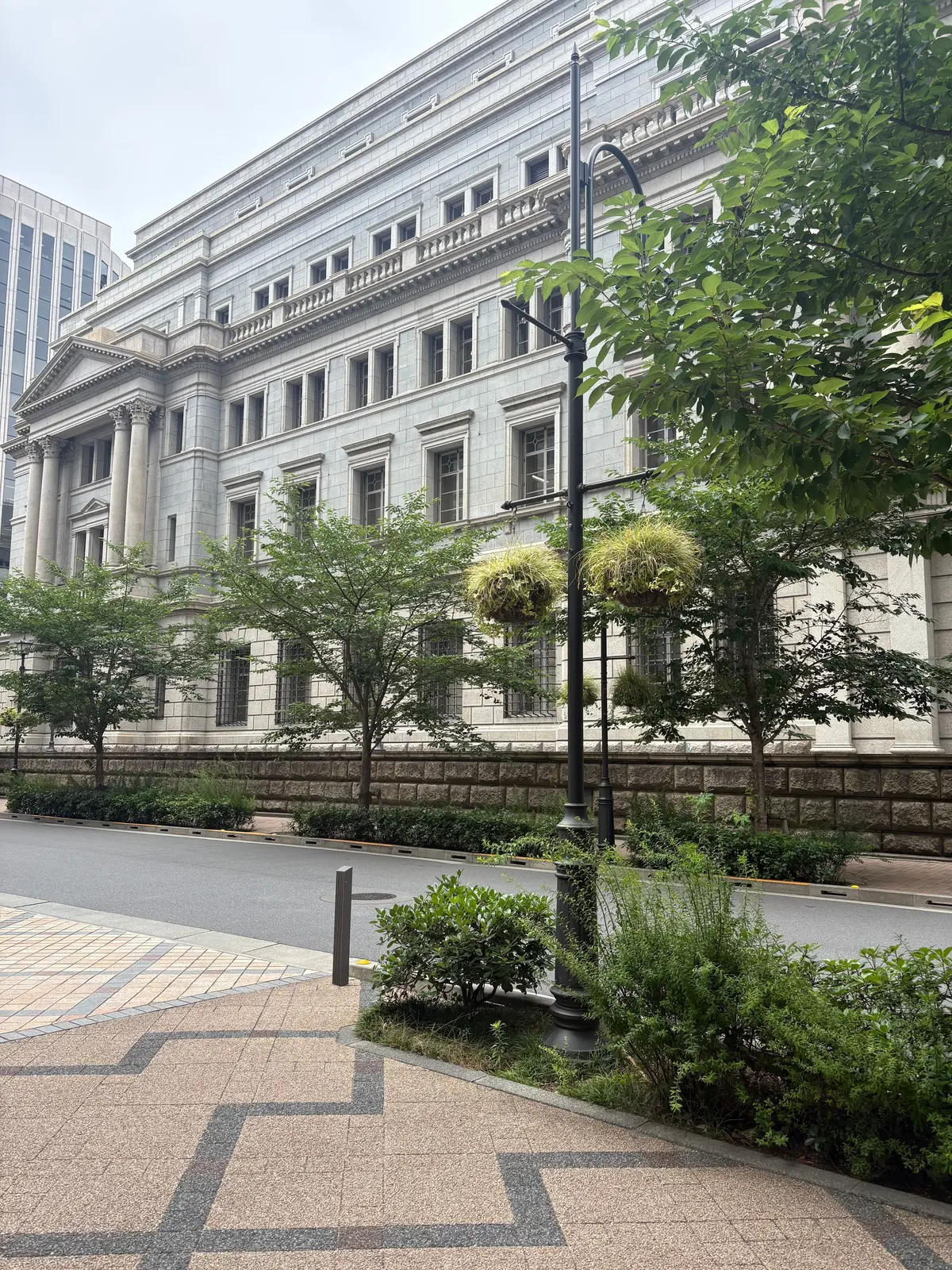 白亜の建物と緑豊かな植栽が調和した風景。正面には古典的な柱を持つ建物、手前には石垣と緑の草木が生い茂り、黒い灯柱が立っている。