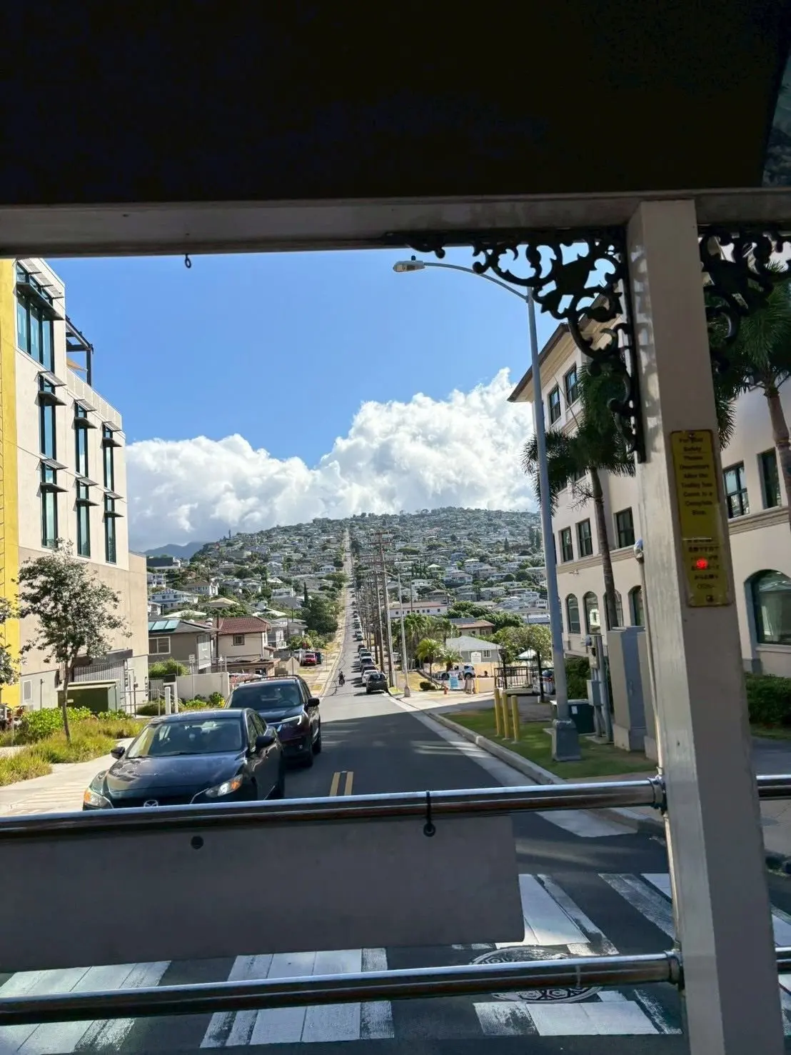 傾斜した道路が山に向かって続き、両脇には建物が並ぶ。青空に白い雲が浮かび、遠景には緑豊かな山並みが見える。手前にバスの窓枠と横断歩道がある。