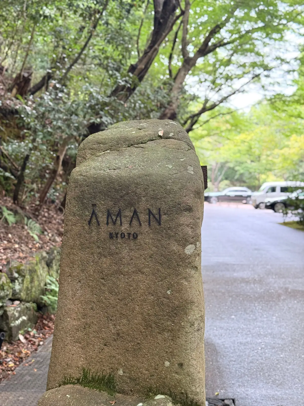 苔むした石柱に「AMAN KYOTO」の文字が刻まれている。背景には緑豊かな木々と駐車場が見える。京都のラグジュアリーリゾートホテル、アマン京都。AMAN京都