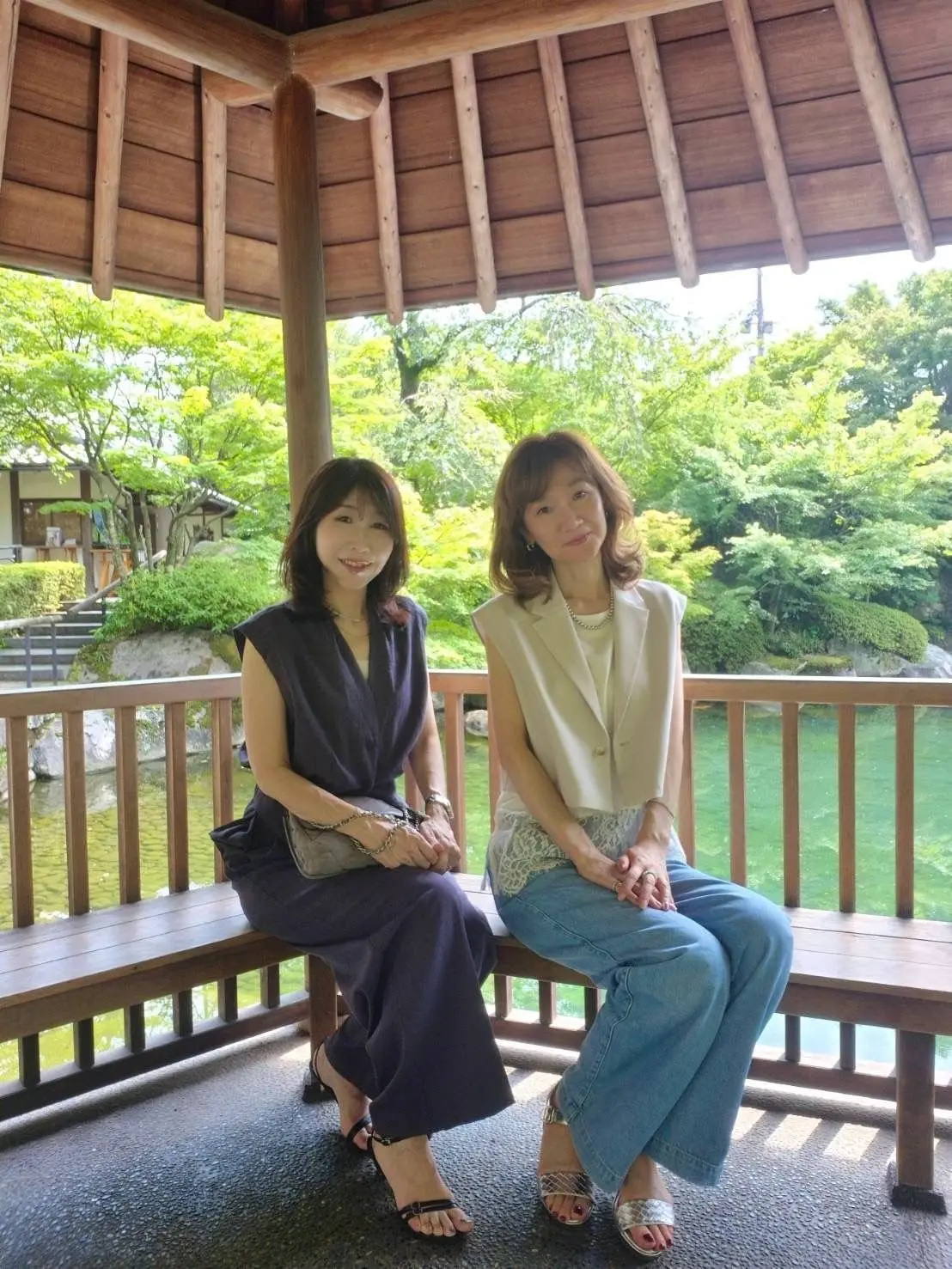 木造の屋根の下、2人の女性がベンチに座っている。左の女性は紺色のノースリーブワンピース、右の女性は白いジャケットとレースのトップス、青いジーンズを着用。背景には緑豊かな庭園と池が見える。