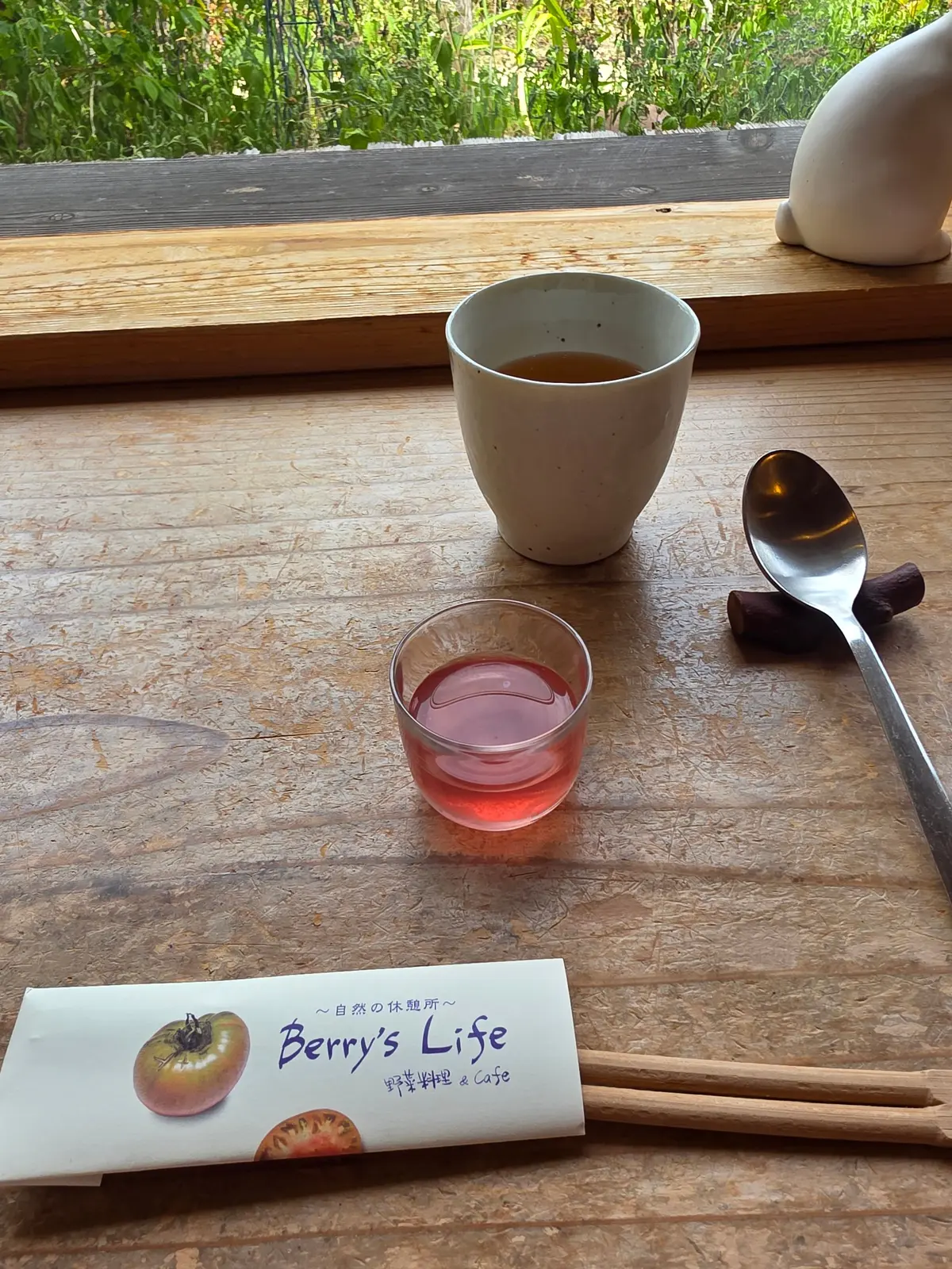 木製のテーブルに置かれた、茶色の飲み物が入った白っぽいカップ、赤いジュースのグラス、スプーン、Berry's Lifeのロゴが入った白い箱。背景には緑の木々が見える。