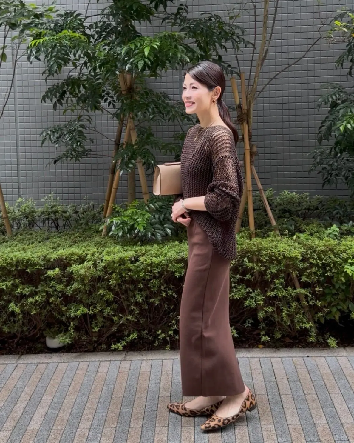 茶色のトップスとロングスカートを着用した女性が、屋外の歩道で歩いている。右手にベージュのハンドバッグを持ち、足元はヒョウ柄のフラットシューズ。背景には緑の植栽とグレーの壁がある。