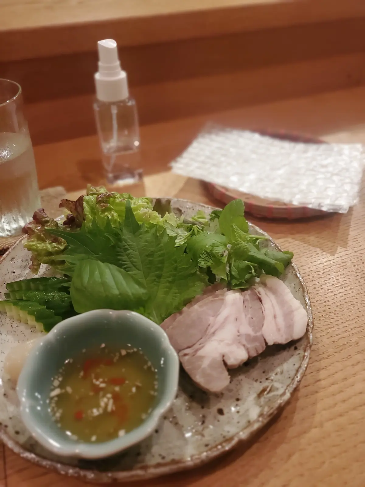 木製のテーブルに置かれた、淡いピンク色の鶏肉と緑色の野菜の盛り合わせ。小皿に盛り付けられ、青い器に入ったドレッシングが添えられている。水を入れたグラスと、白いスプレーボトル、ナプキンも写っている。