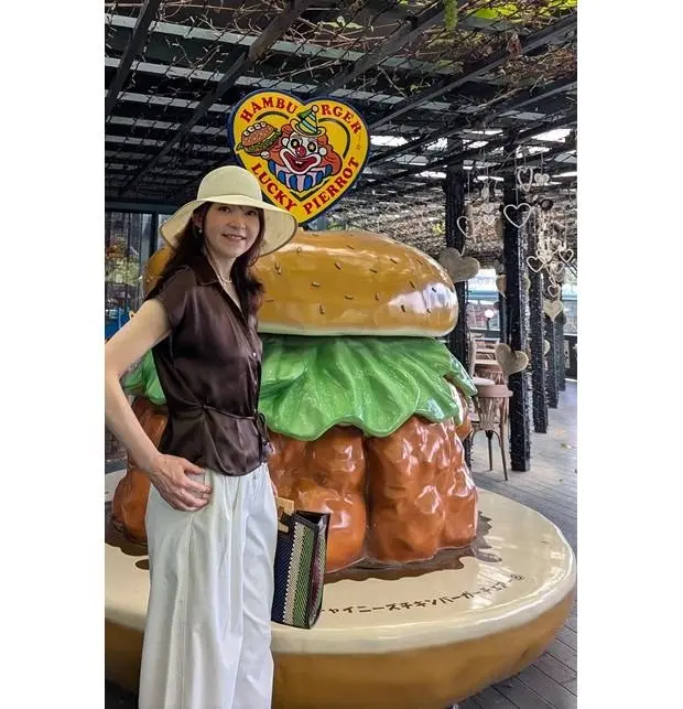 ハットを被った女性が、巨大なハンバーガーのオブジェの横でポーズをとっている。背景には「HAMBURGER LUCKY PIERROT」のロゴがある。