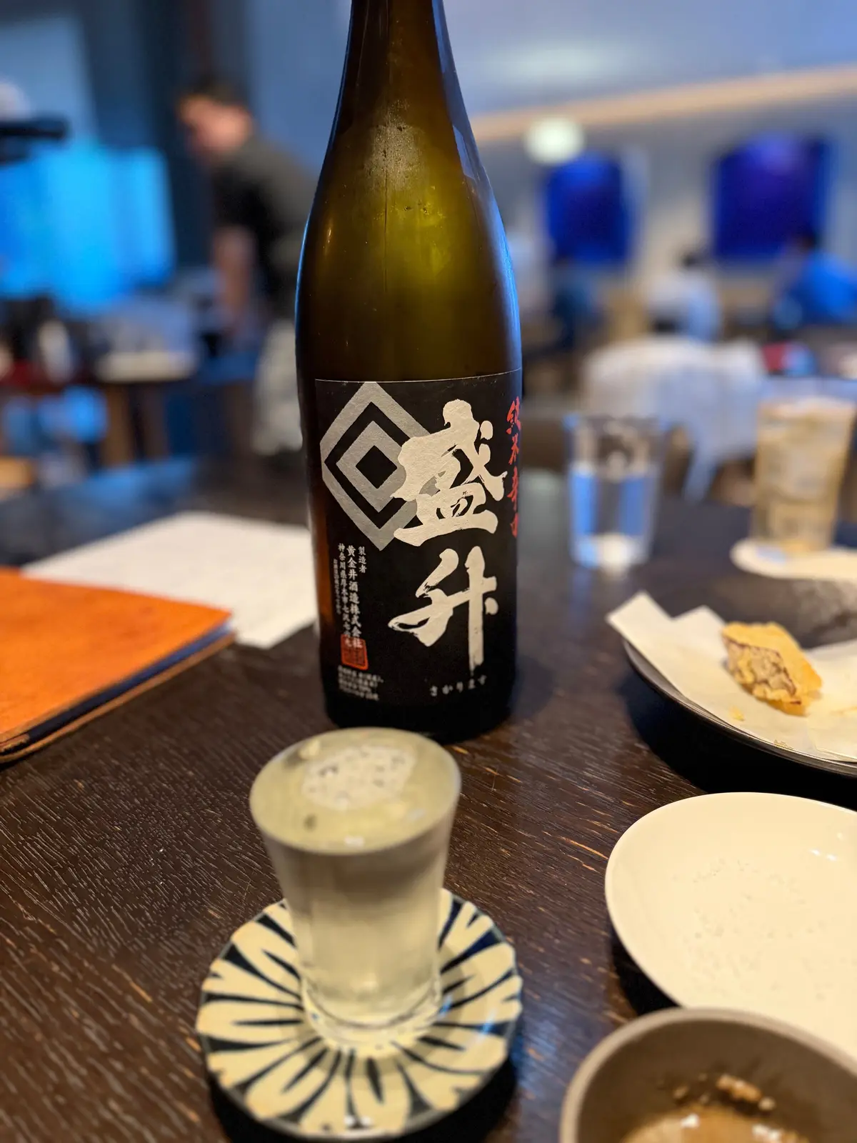 ガラスの杯に注がれた日本酒と、黒ラベル付きのボトルが木製のテーブルに置かれている。テーブルには白い食器や小皿も並んでいる。