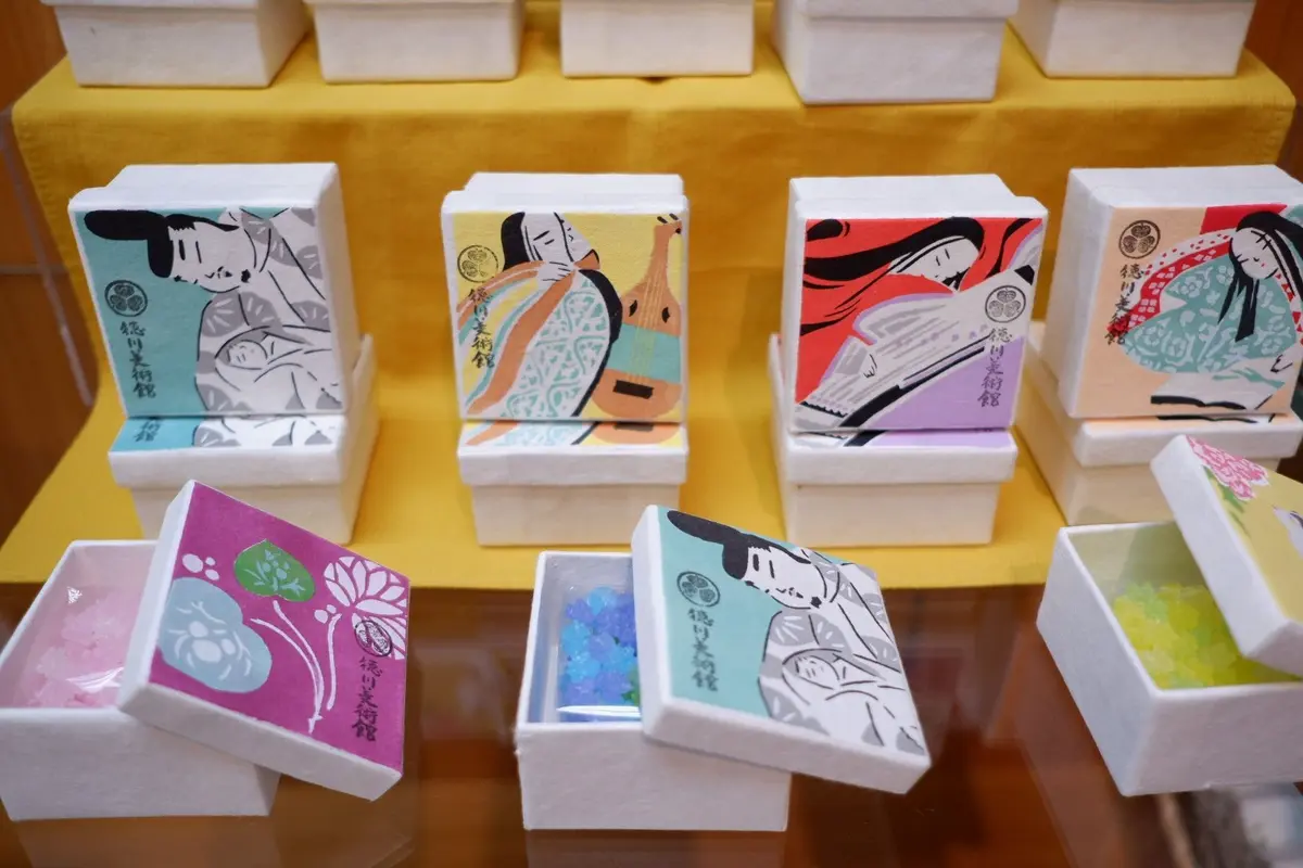 黄色のテーブルに並んだ白い箱で、伝統的な日本風の絵が描かれている。箱の一部が開いていて、ピンクや青の小さな商品が入っている。