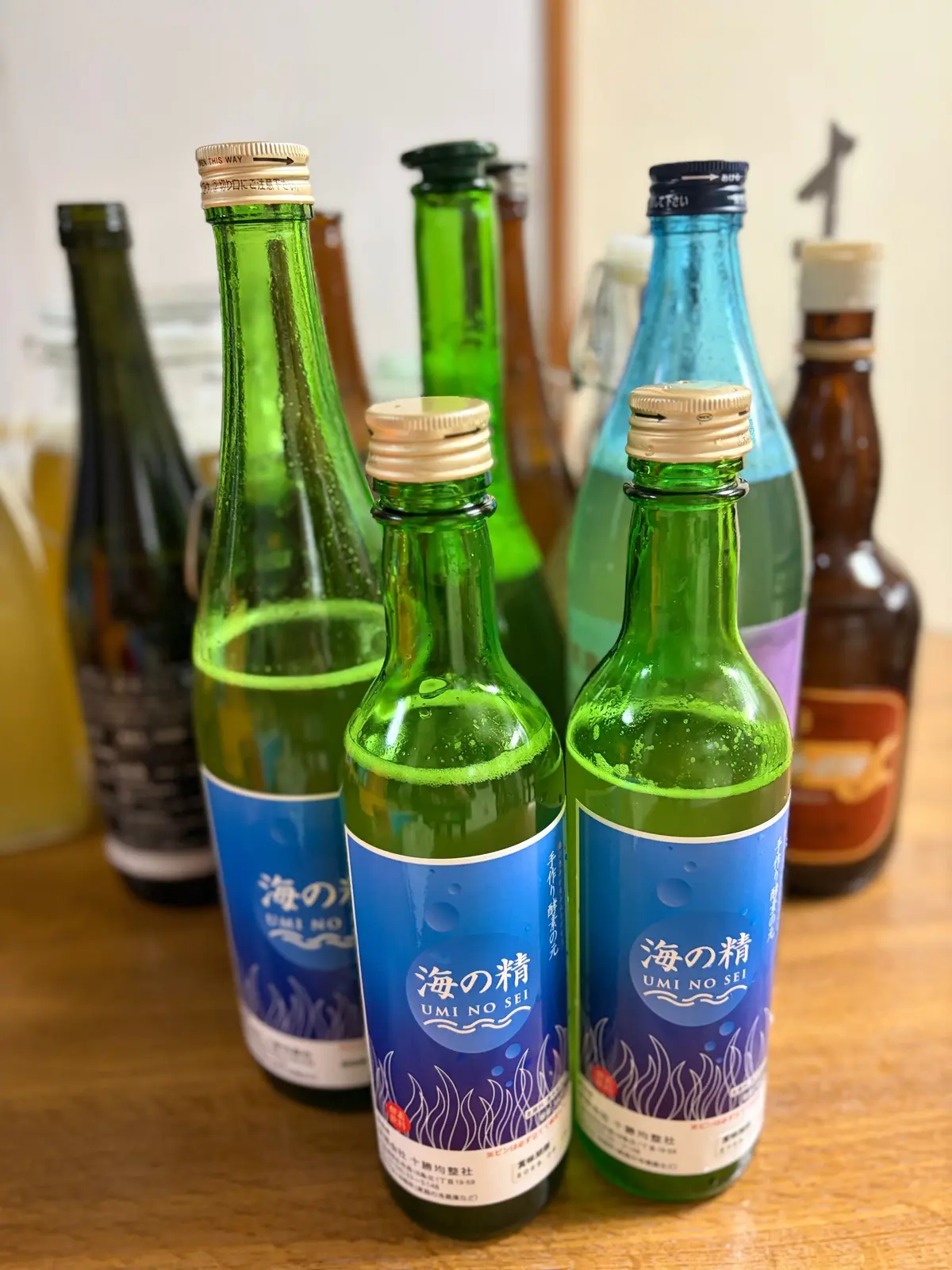 緑色のガラス瓶が複数並び、青いラベルと日本語の文字が目立つ。中央に「海の醸」のラベルが2本あり、背景に他の瓶も少し見える。