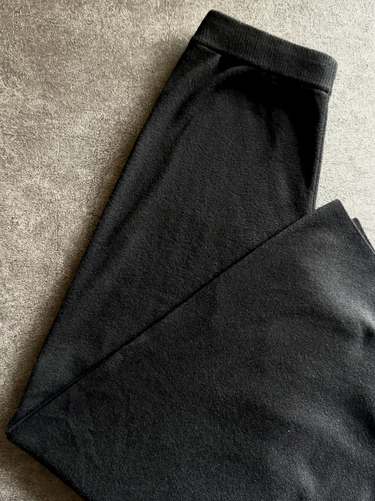 黒いスウェットパンツが畳まれて置かれている。素材は起毛で、全体的にマットな質感。左から右に傾いた状態で、裾が少し見える。