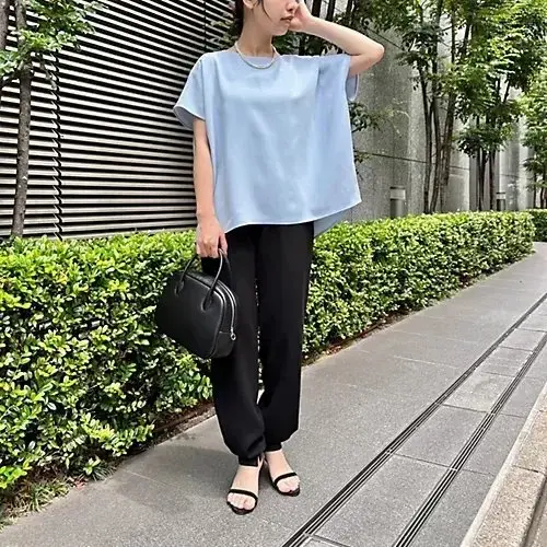 青色のゆったりとしたトップスに黒いパンツを合わせた女性。右手に黒いハンドバッグを持ち、左手で頭を支えている。背景には緑の植栽とコンクリートの建物がある。