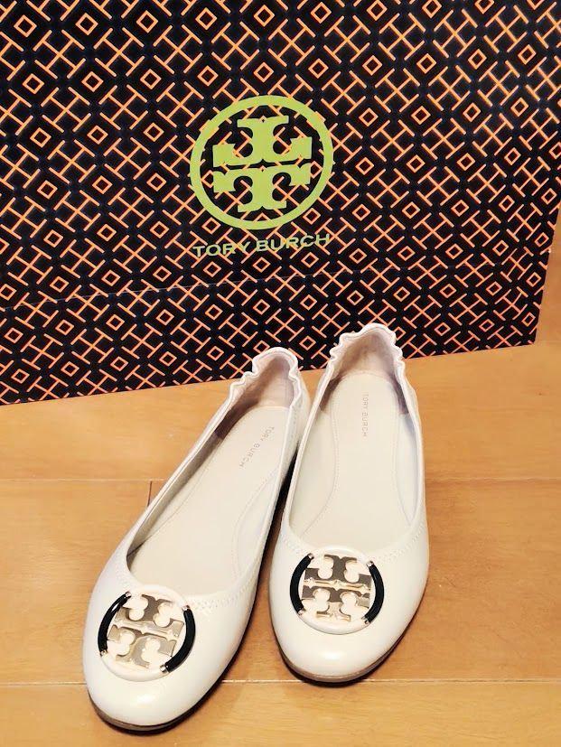 白の Tory Burch（トリーバーチ）フラットシューズが2つ並び、背景にはブランドの箱が置かれている。箱にはオレンジ色のロゴがデザインされている。