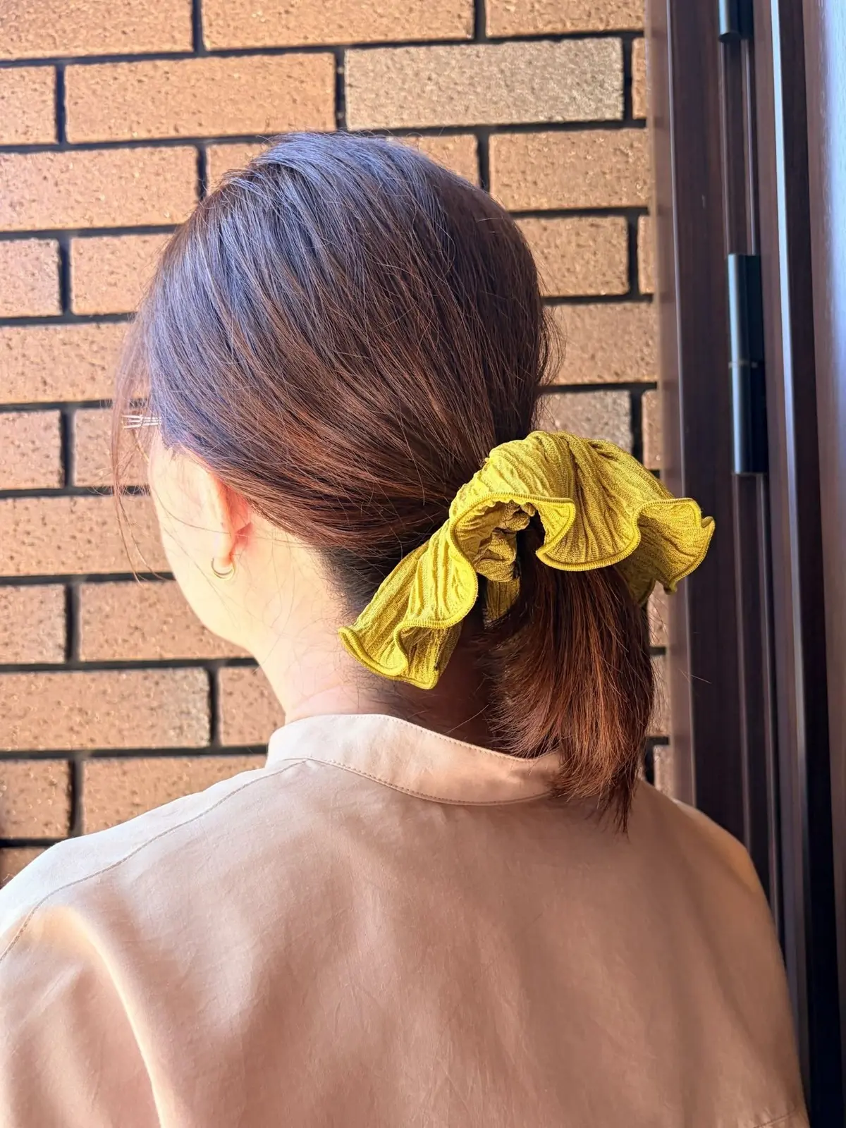 茶色の髪を後ろで一つに束ねた女性。黄色のフリル付きヘアゴムで飾っている。背景にはレンガの壁とドアがある。