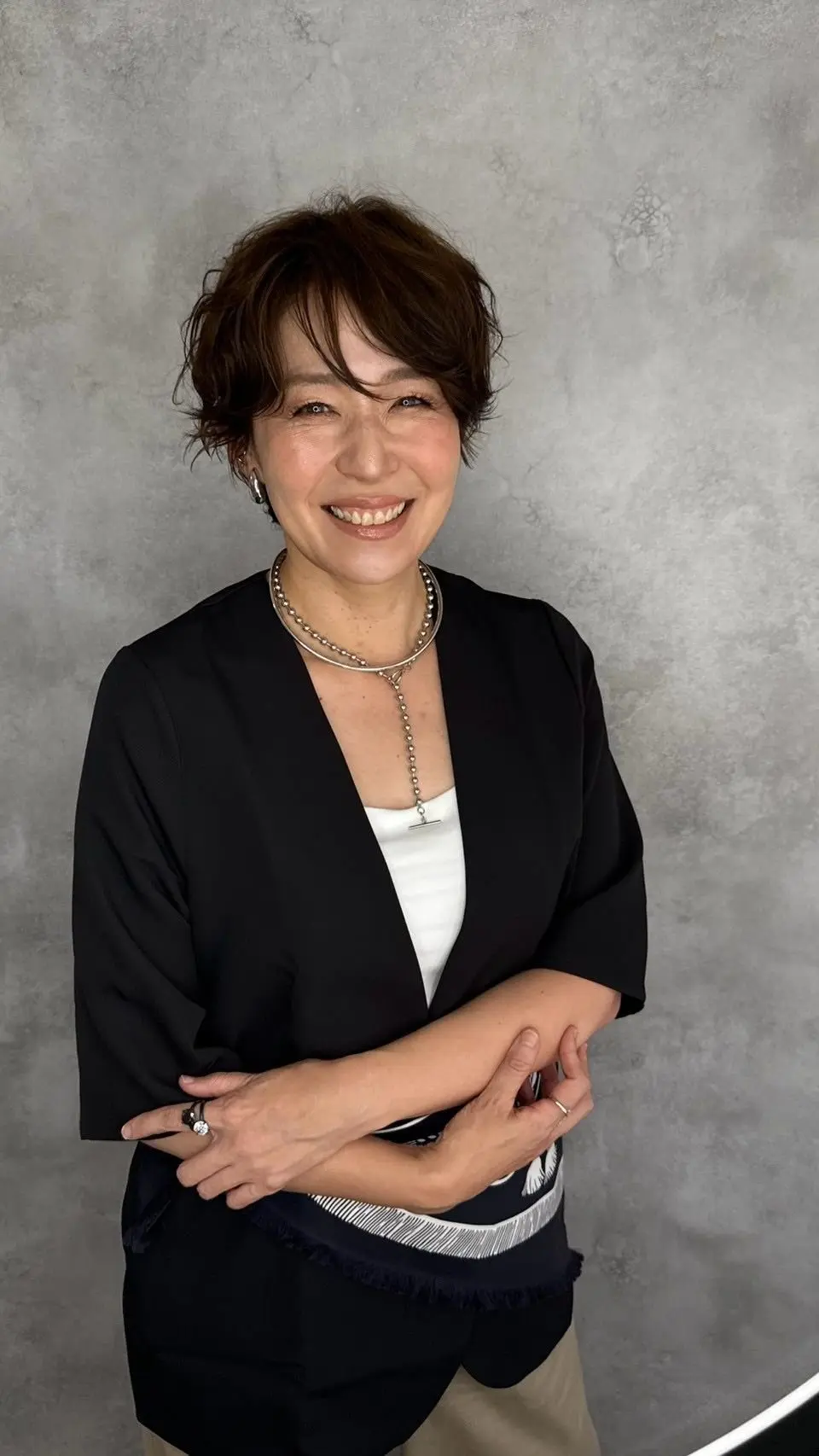 笑顔の女性が、黒のジャケットと白いインナーを着用して立っているポートレート。茶色の短髪で、シルバーのネックレスを身につけている。背景はグレーのテクスチャードな壁。