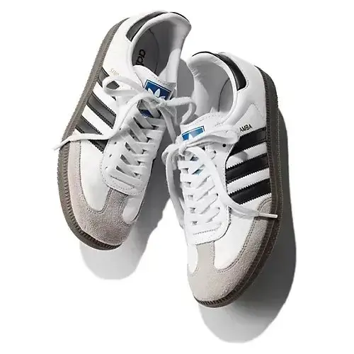 白とグレーのAdidas Sambaスニーカーが左右一対、白い背景に置かれている。黒の三本線が側面にあり、白いシューレースが通っている。