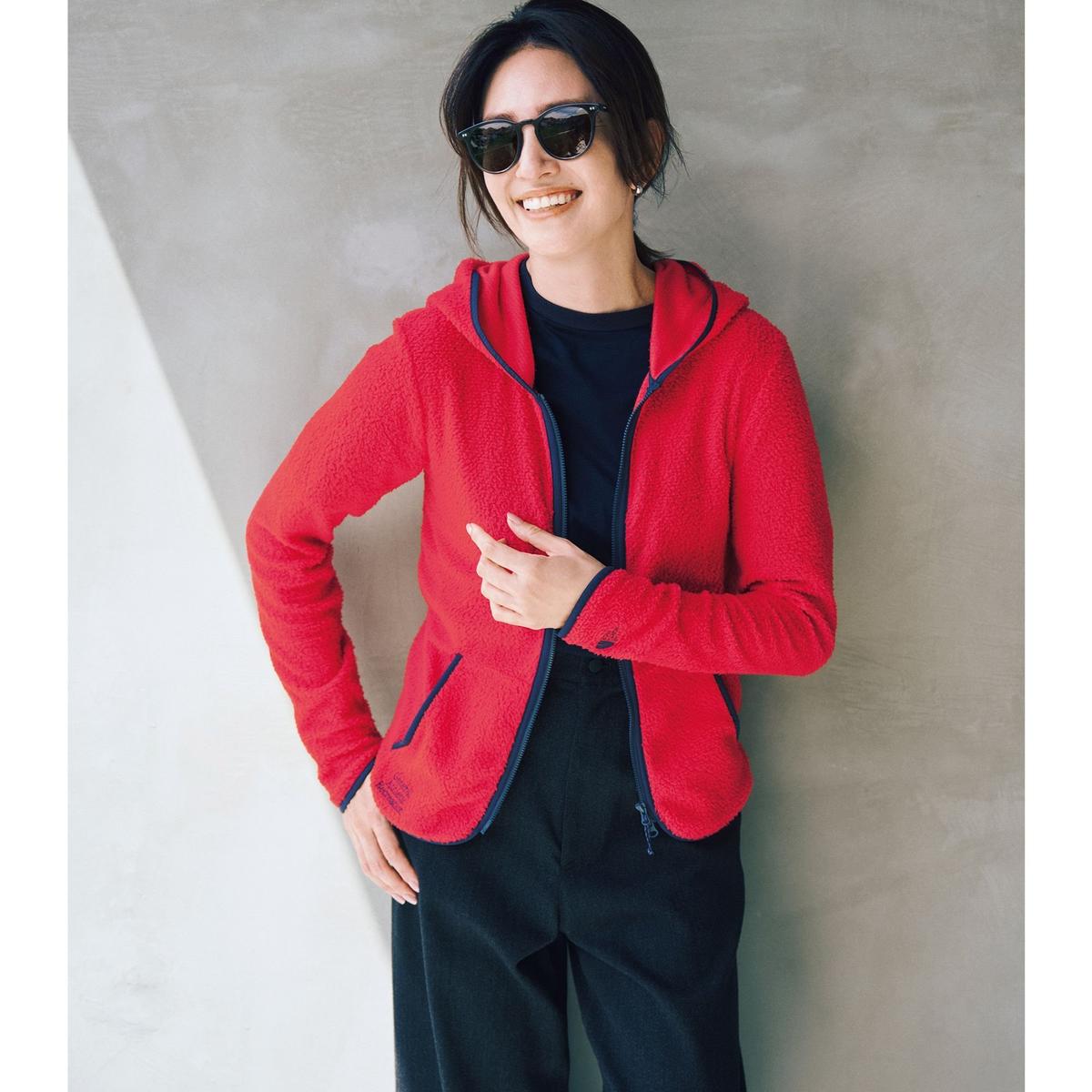赤いフリース素材のパーカーを着た女性。黒いカットソーの上に着用し、黒いパンツを合わせたカジュアルな装い。サングラスをかけ、微笑んでいる。背景はコンクリートの壁。