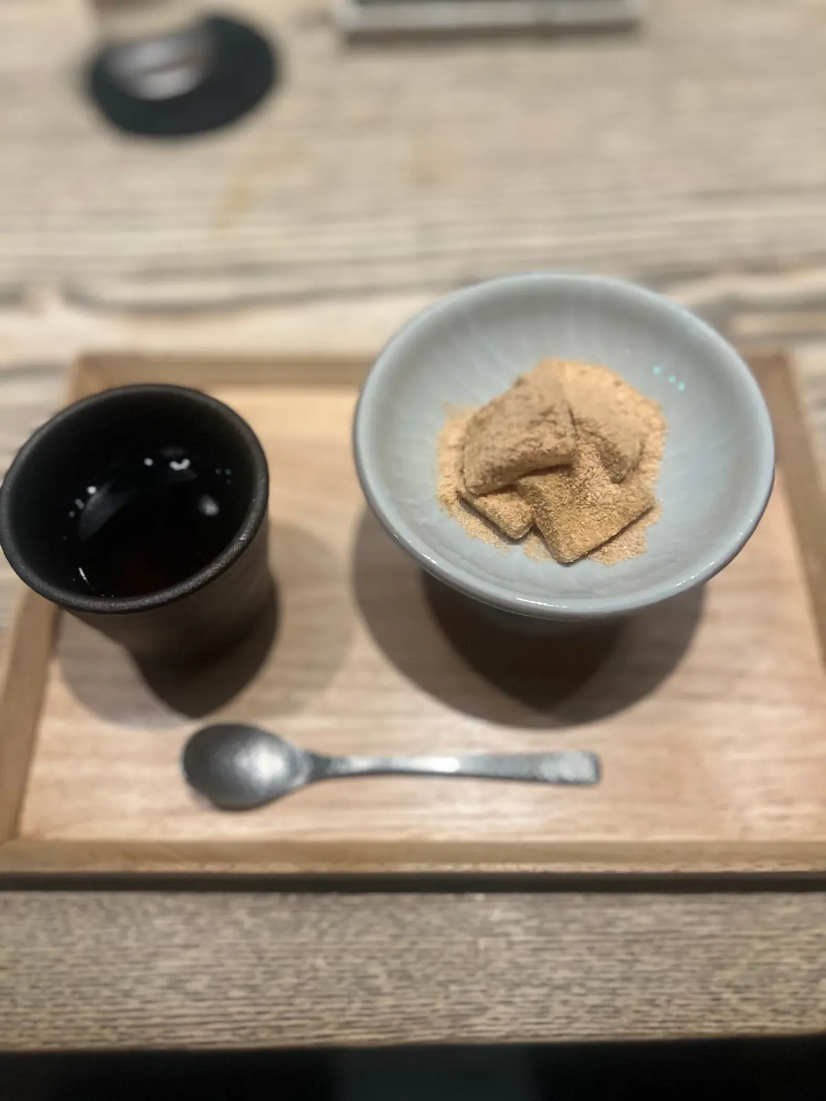 木製トレイに、黒いカップに注がれた濃い液体、小さな白いボウルに積み重ねられた茶色い角形の菓子、銀のスプーンが置かれている。